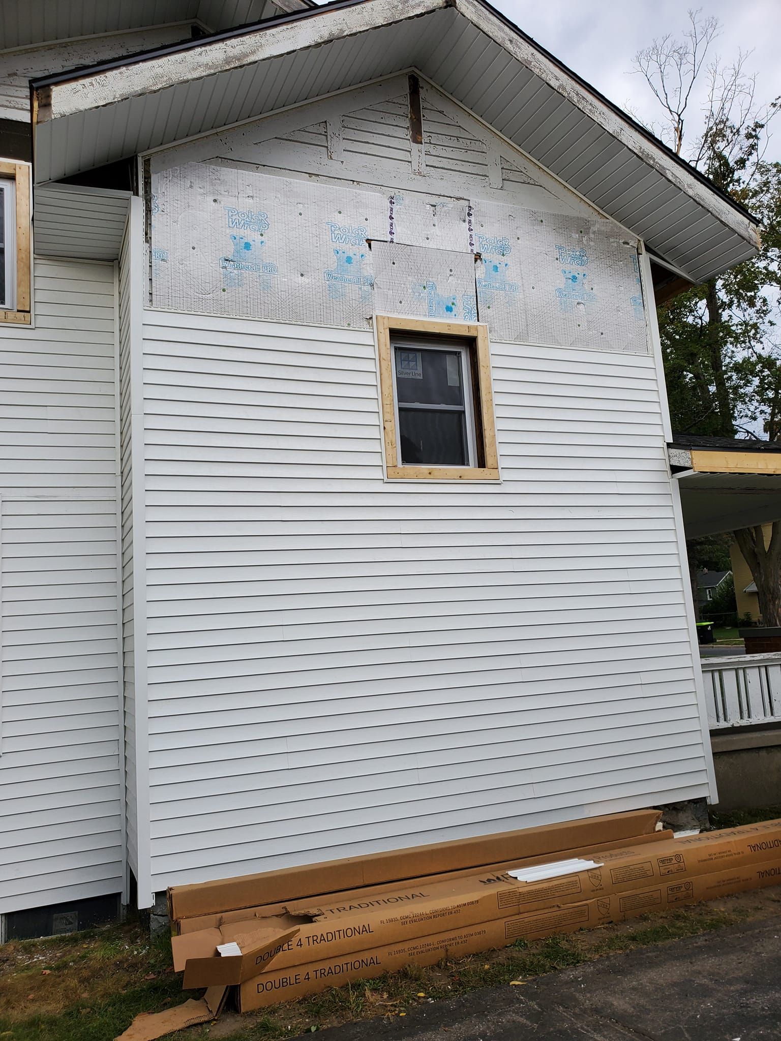 Side view of a house with white siding, damaged upper wall, and a small window above stacked lumber.