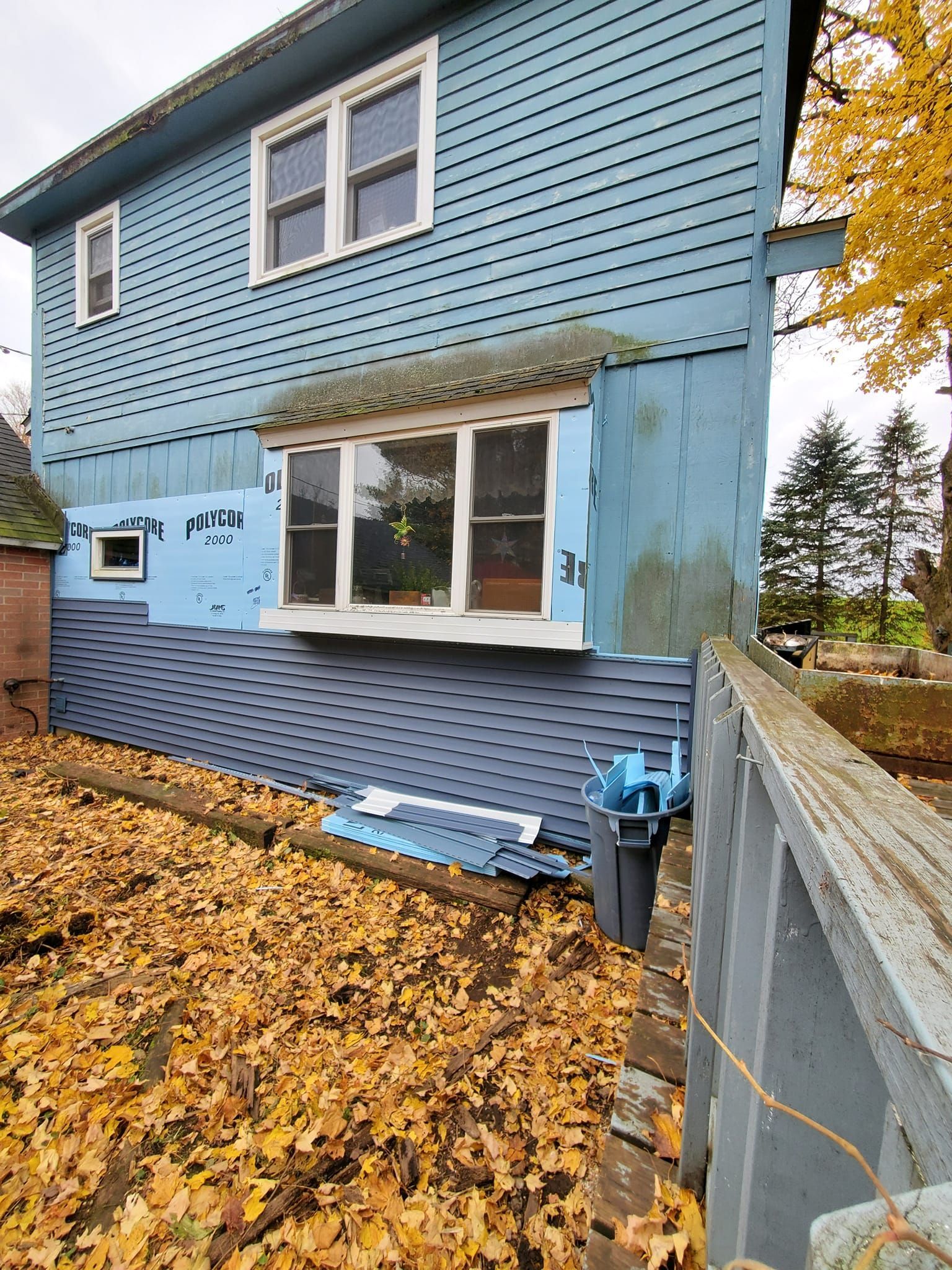 Blue two-story house with leaf-covered yard and fence in autumn