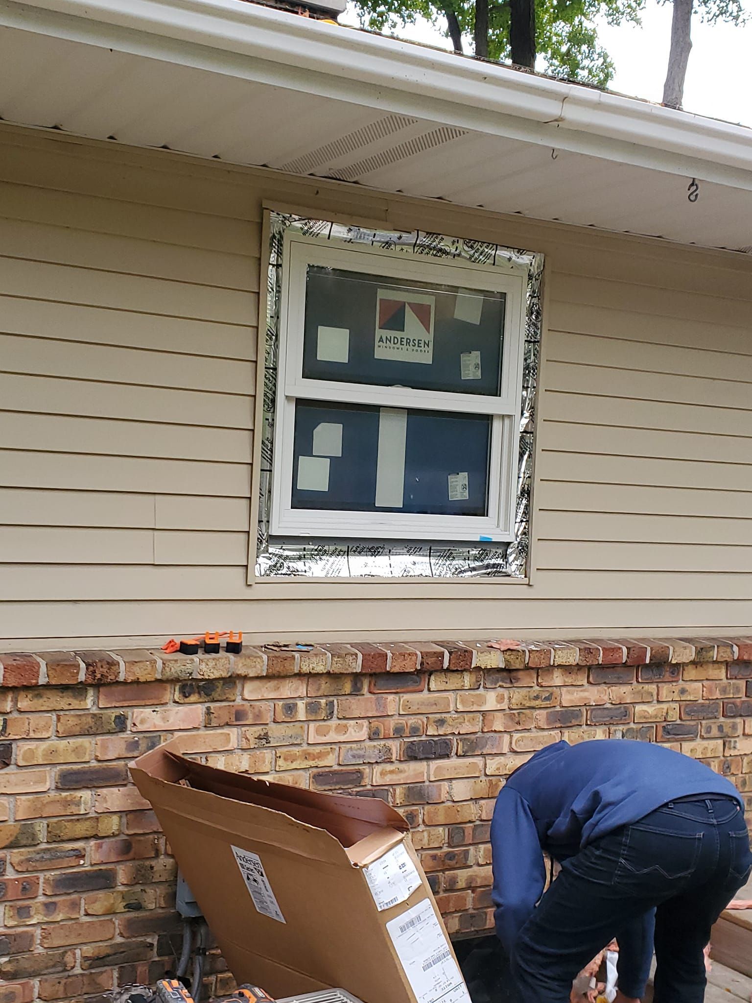Person working near a house window with a cardboard box and brick siding