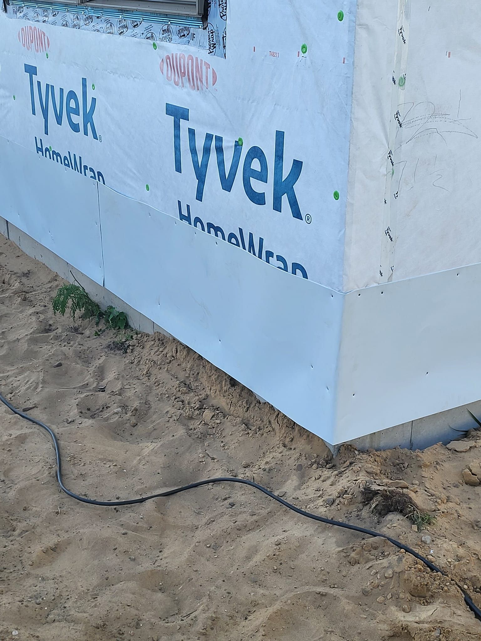 Tyvek house wrap on exterior wall corner above sandy ground with black cable