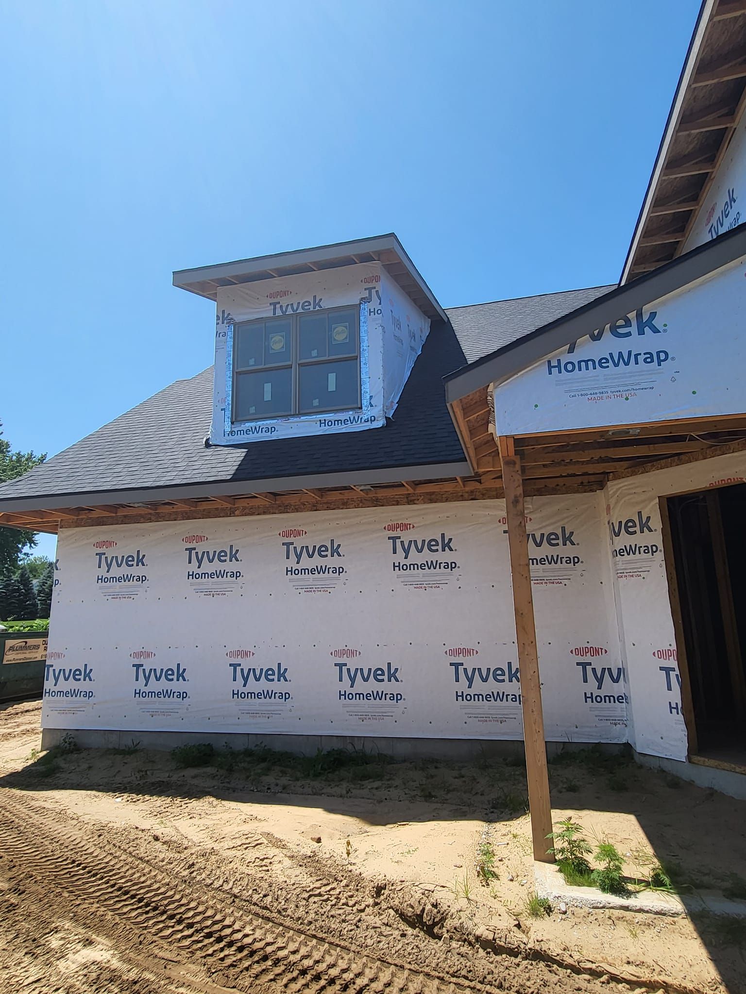 House exterior under construction with blue roof, dormer window, and wrapped walls in bright sunlight