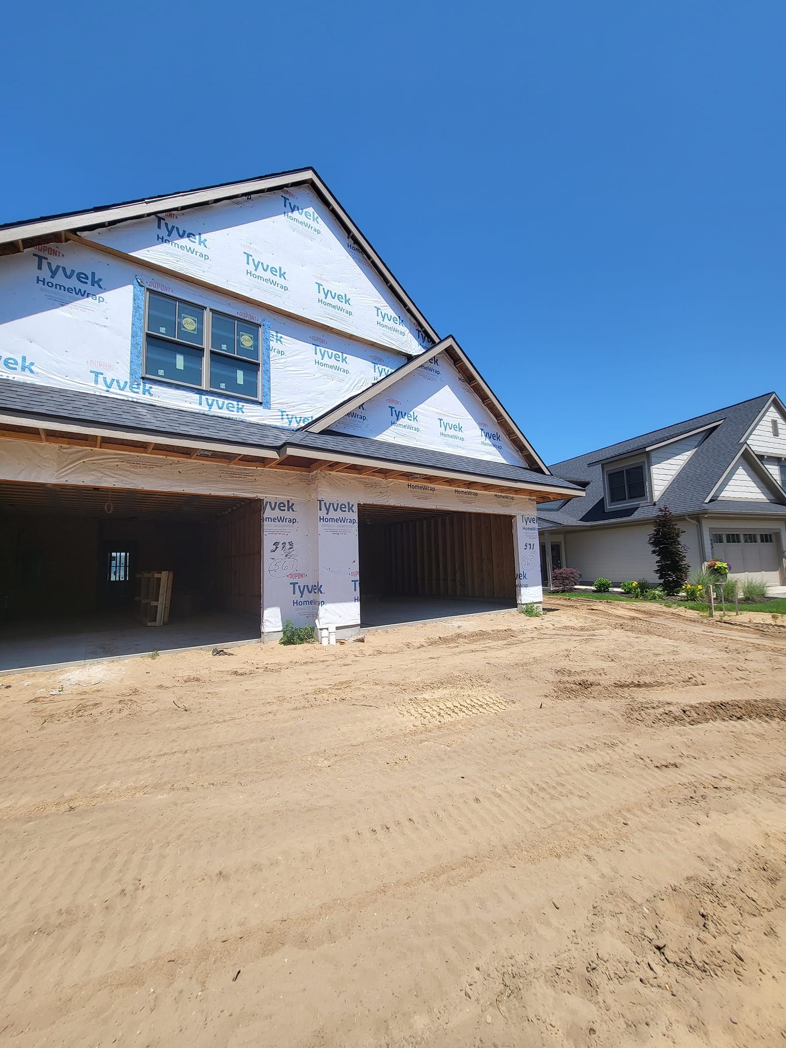 New house under construction with wrapped exterior and dirt driveway under a clear blue sky