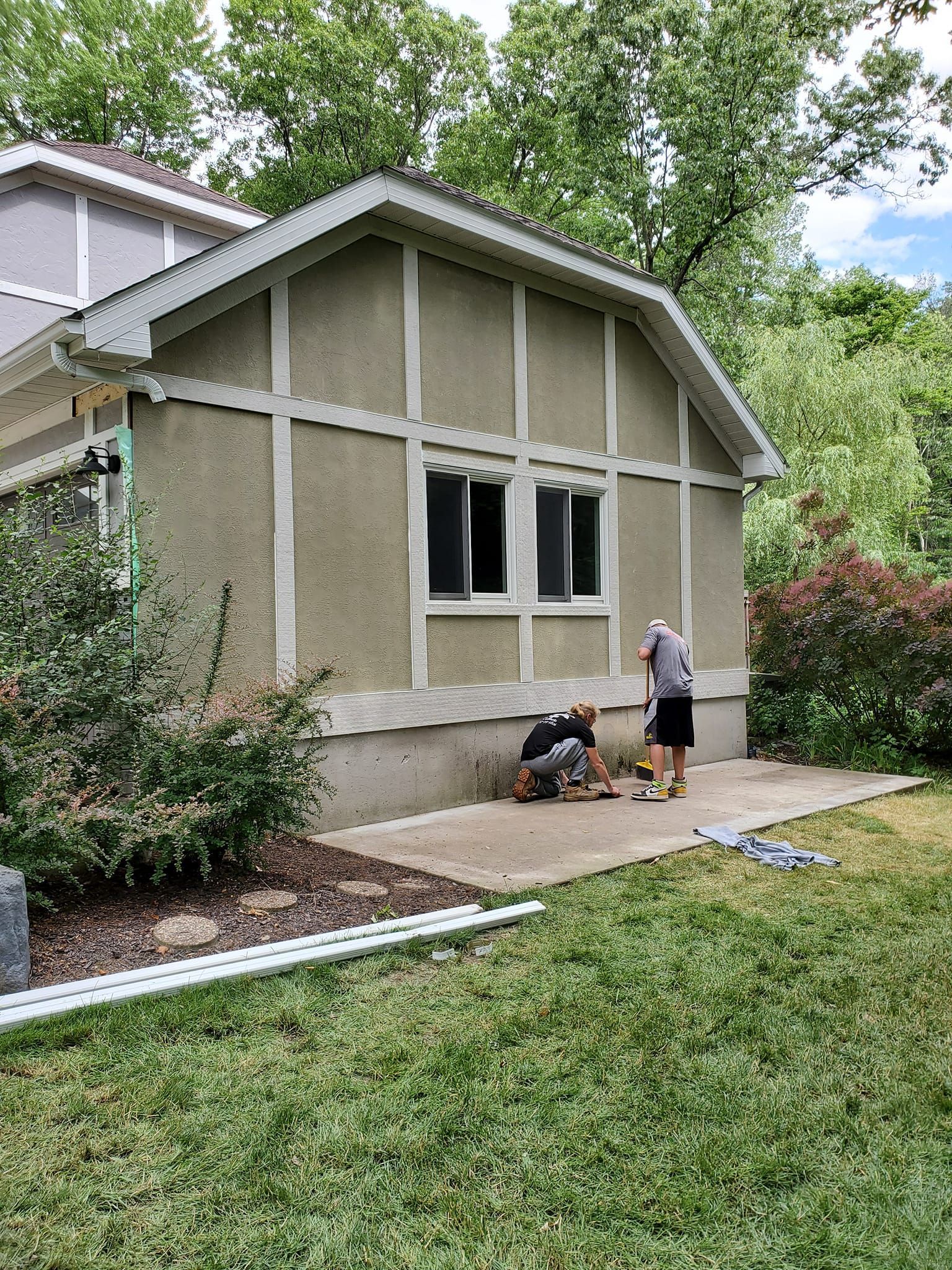 Two people working on the front of a beige house, with siding and trim being repaired.