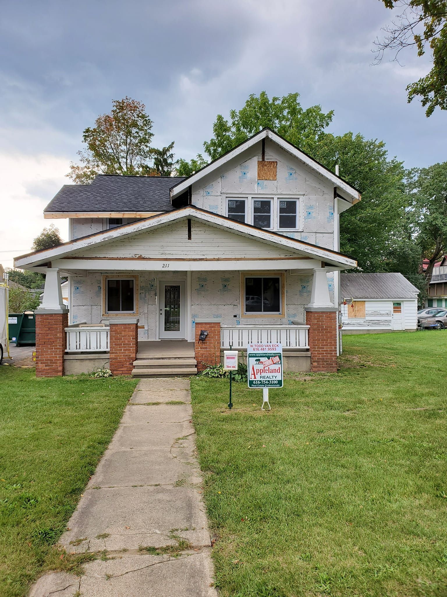 Small white house with a front porch, brick columns, and a lawn sign, viewed from the sidewalk.