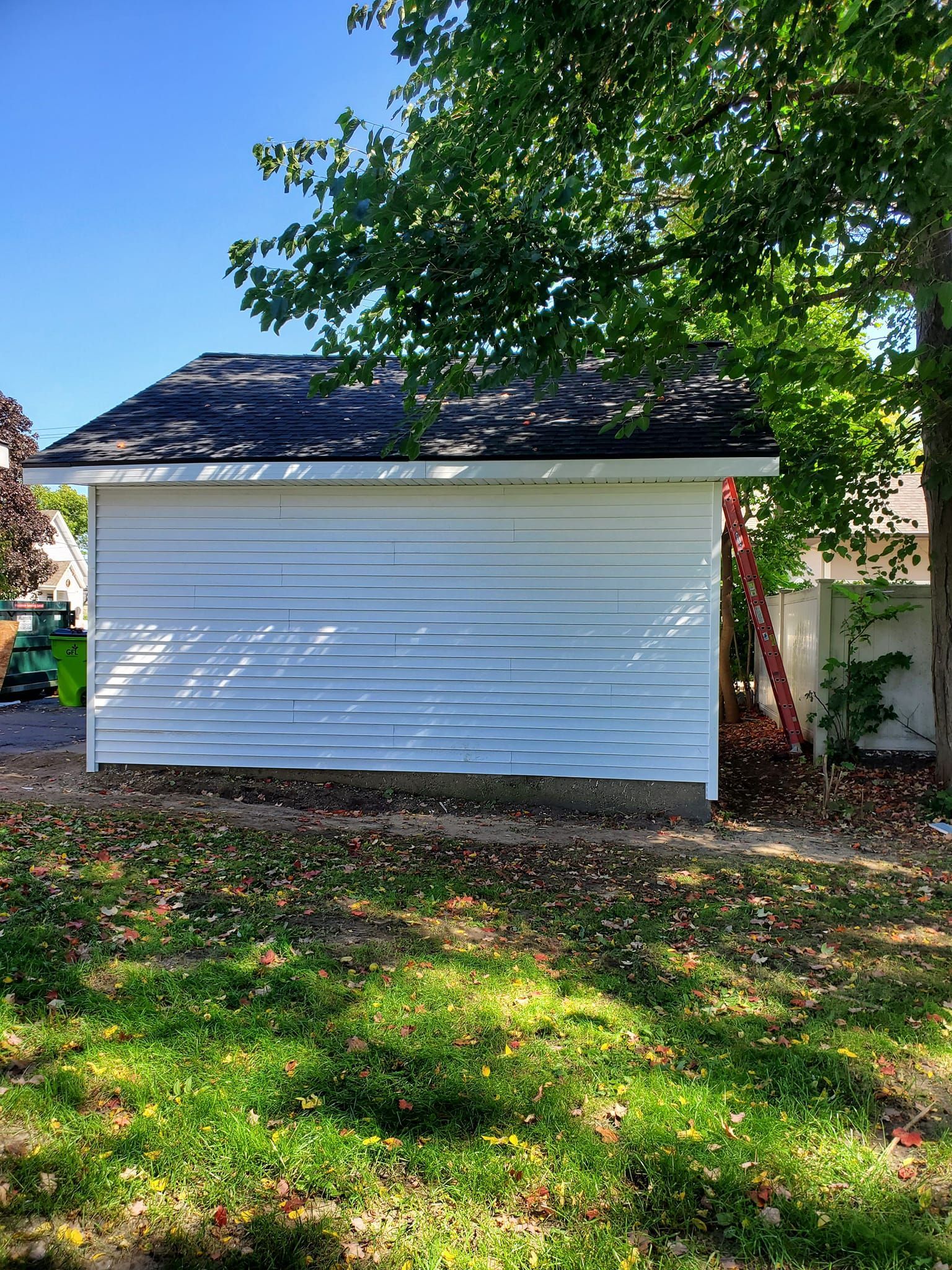 White detached garage with dark roof beside a leafy tree and grassy yard on a sunny day
