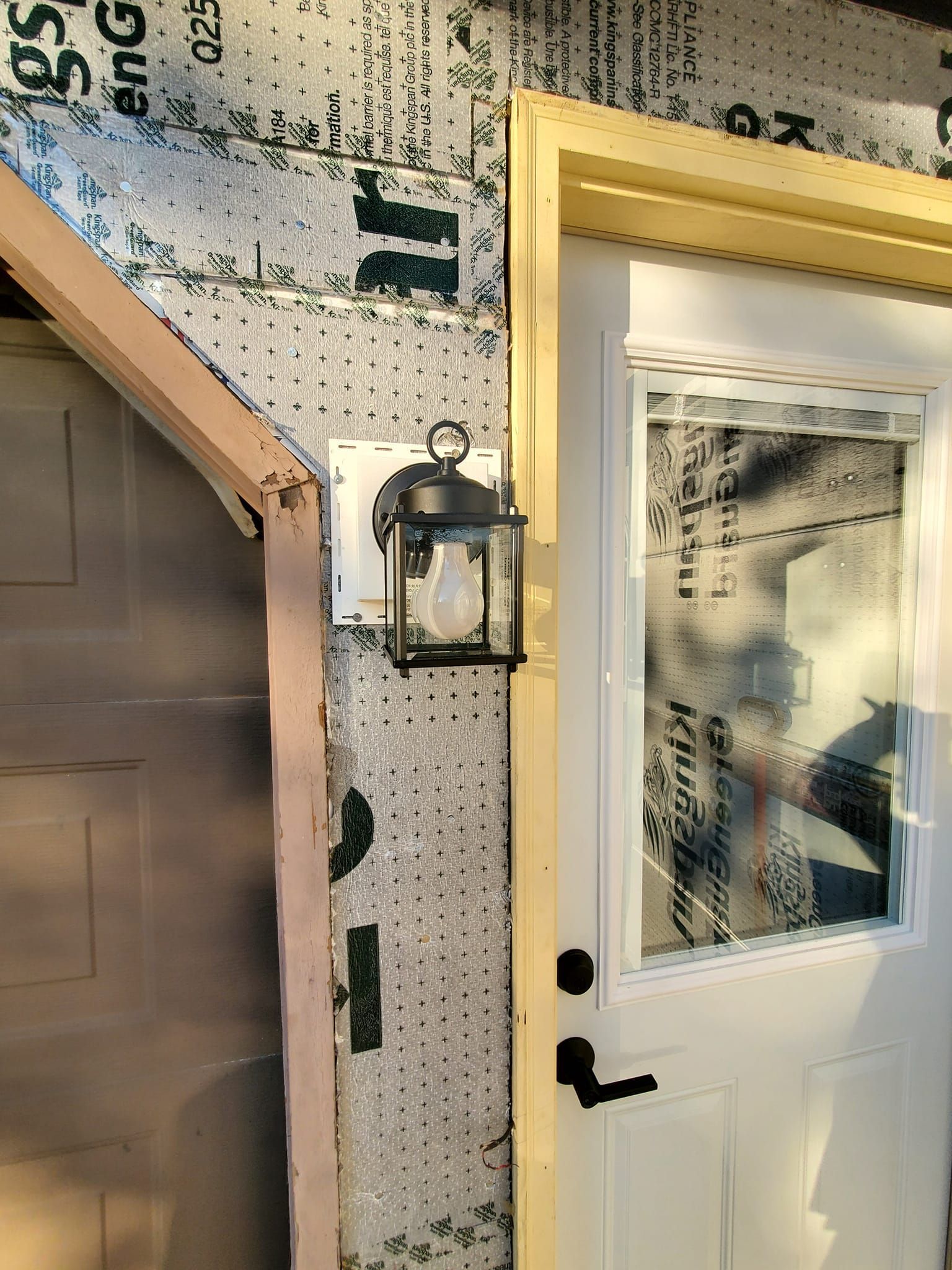 Exterior of a house with a white door, wall lantern, and gray garage door under construction wrap