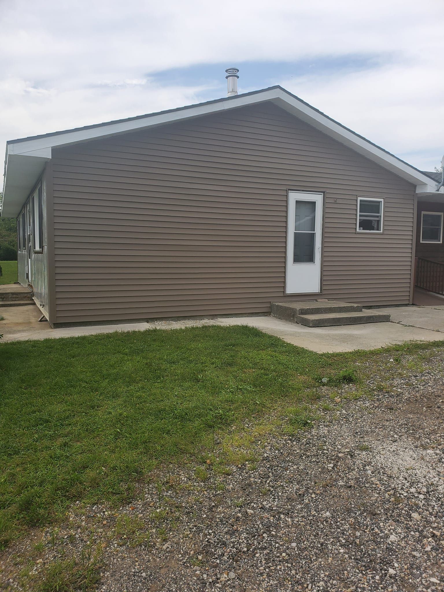 Tan house exterior with white door, gravel driveway, and green lawn under an overcast sky