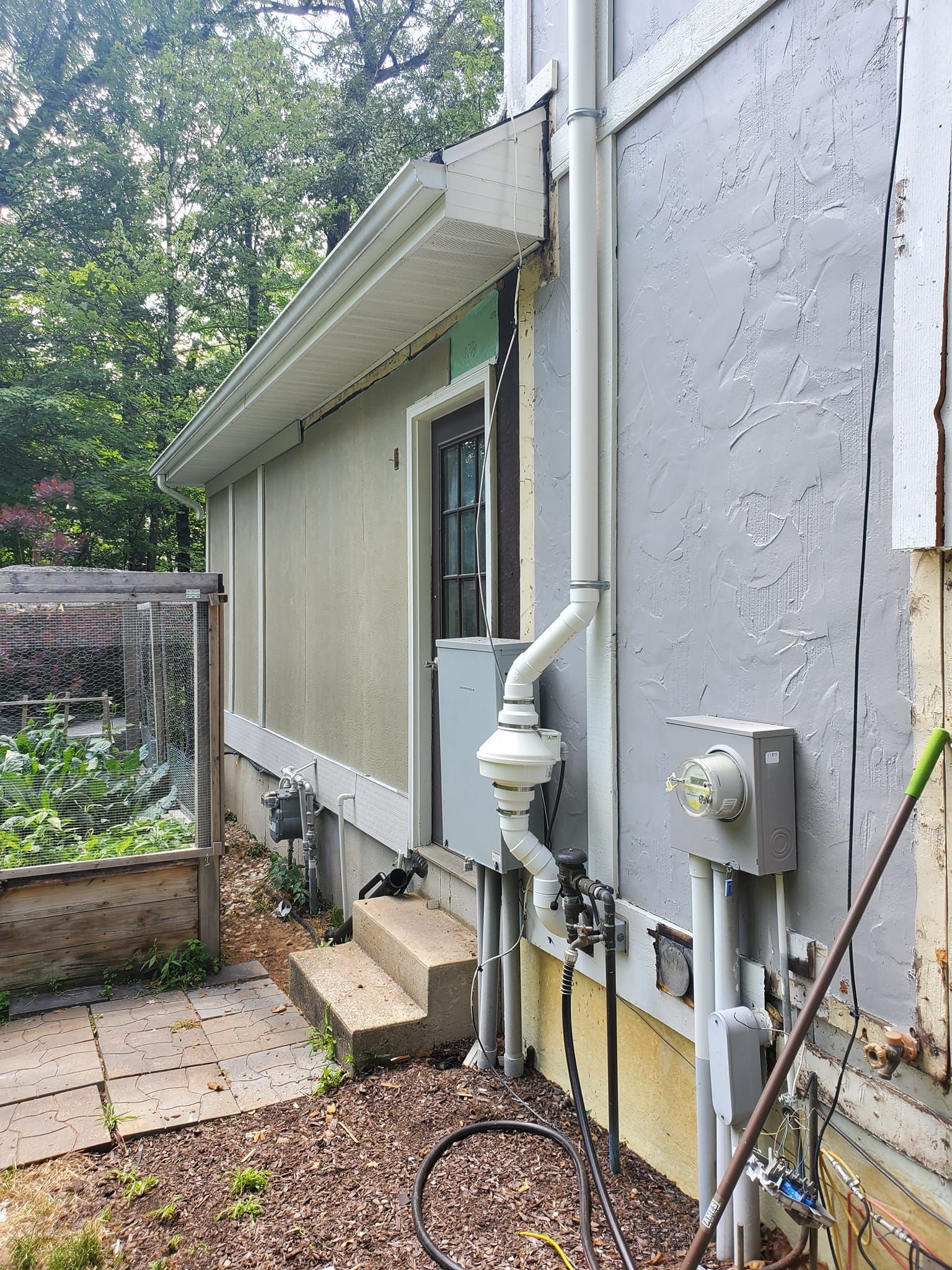 Side view of a house exterior with gray siding, utility meter, pipes, and a small gravel walkway next to a fence.