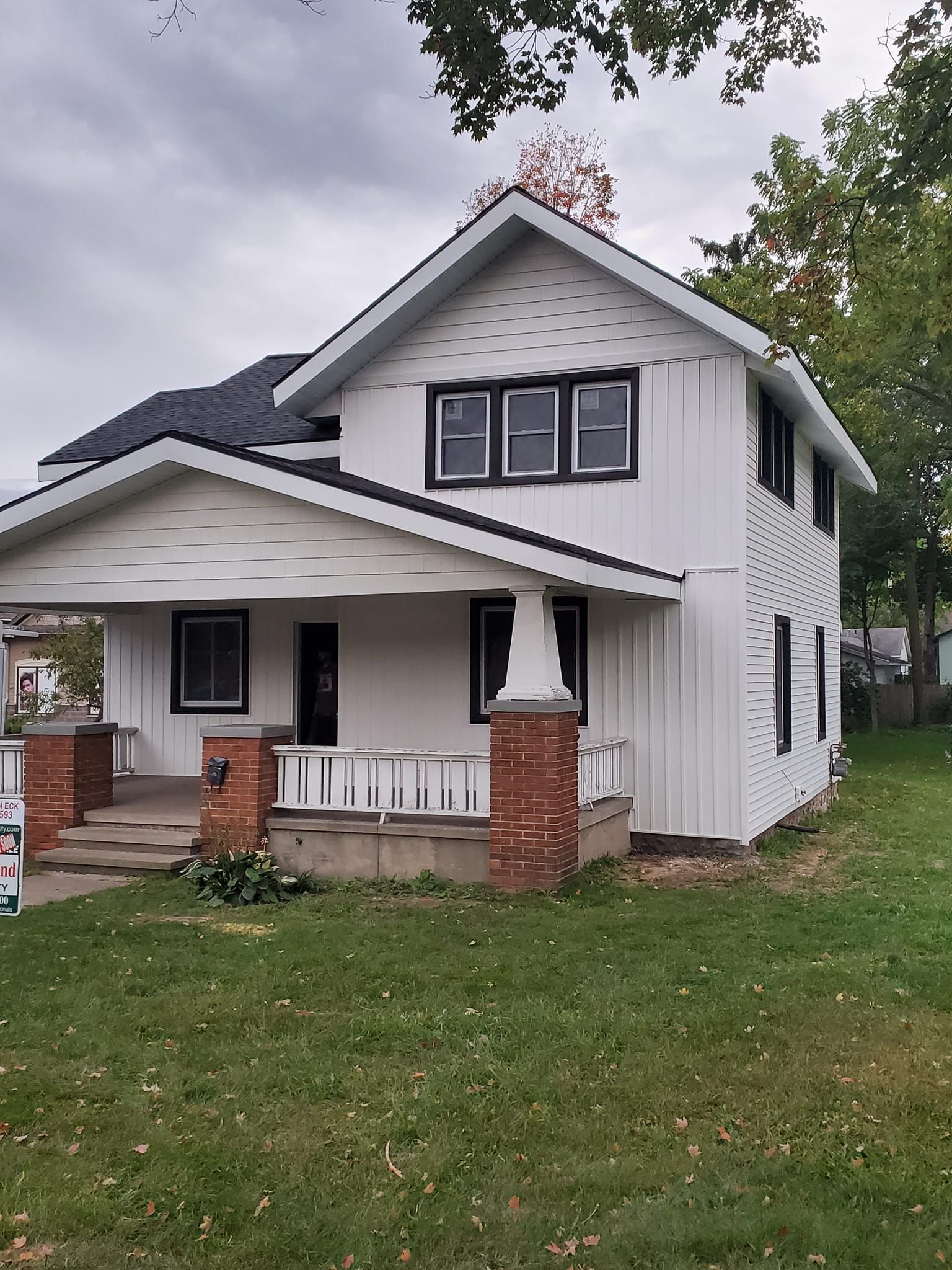 Small white house with black trim, front porch, and brick columns on a grassy lot under cloudy skies