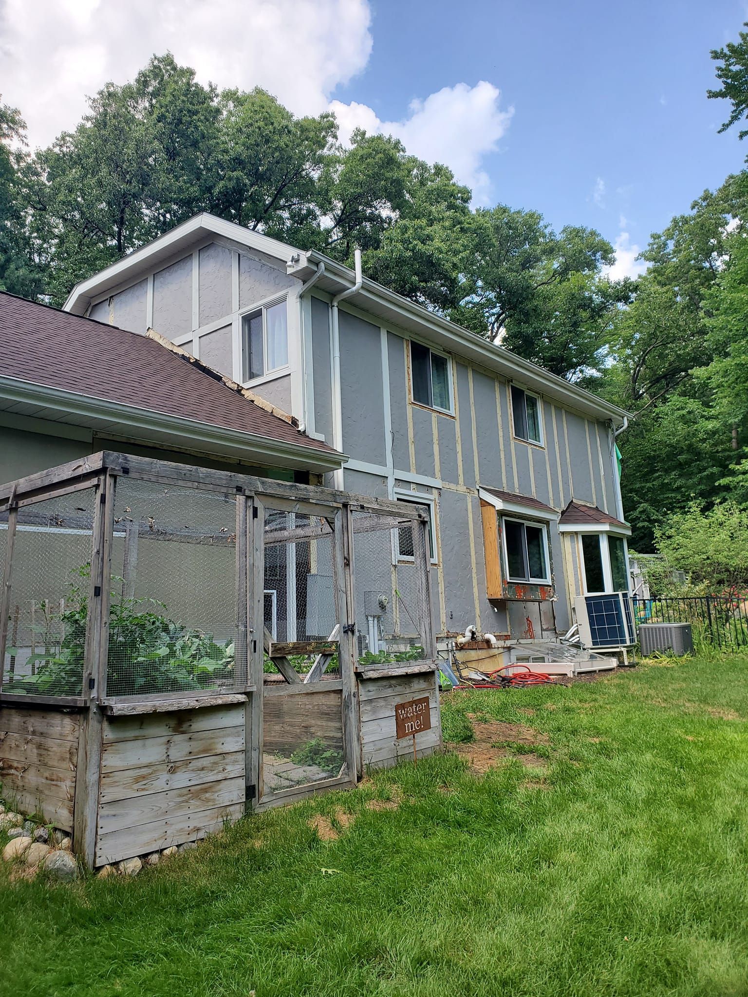 Gray two-story house with a screened porch and fenced garden in a grassy yard under a blue sky