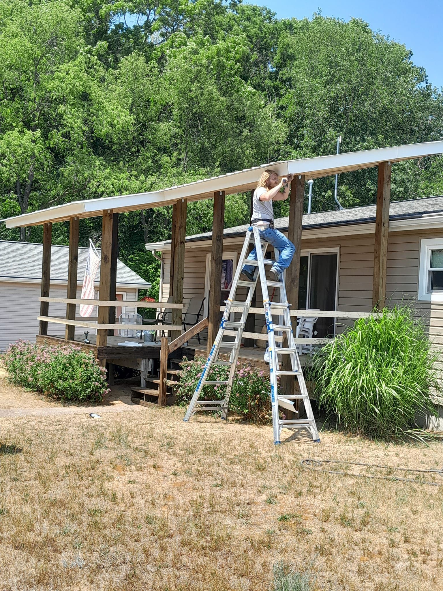 Person on a ladder repairing a roof over a small porch beside a house.