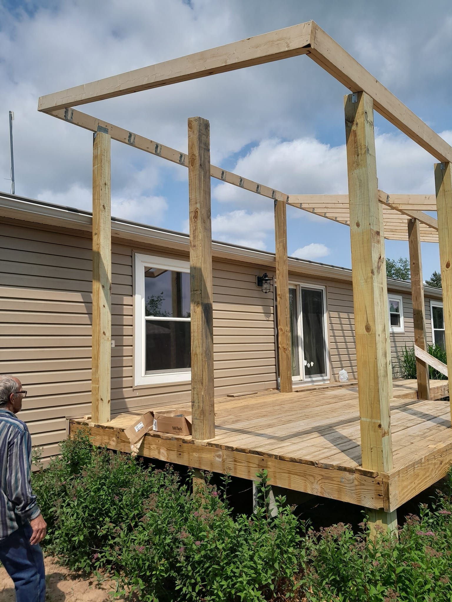 Wooden deck frame under construction beside a house, with vertical posts and roof beams.
