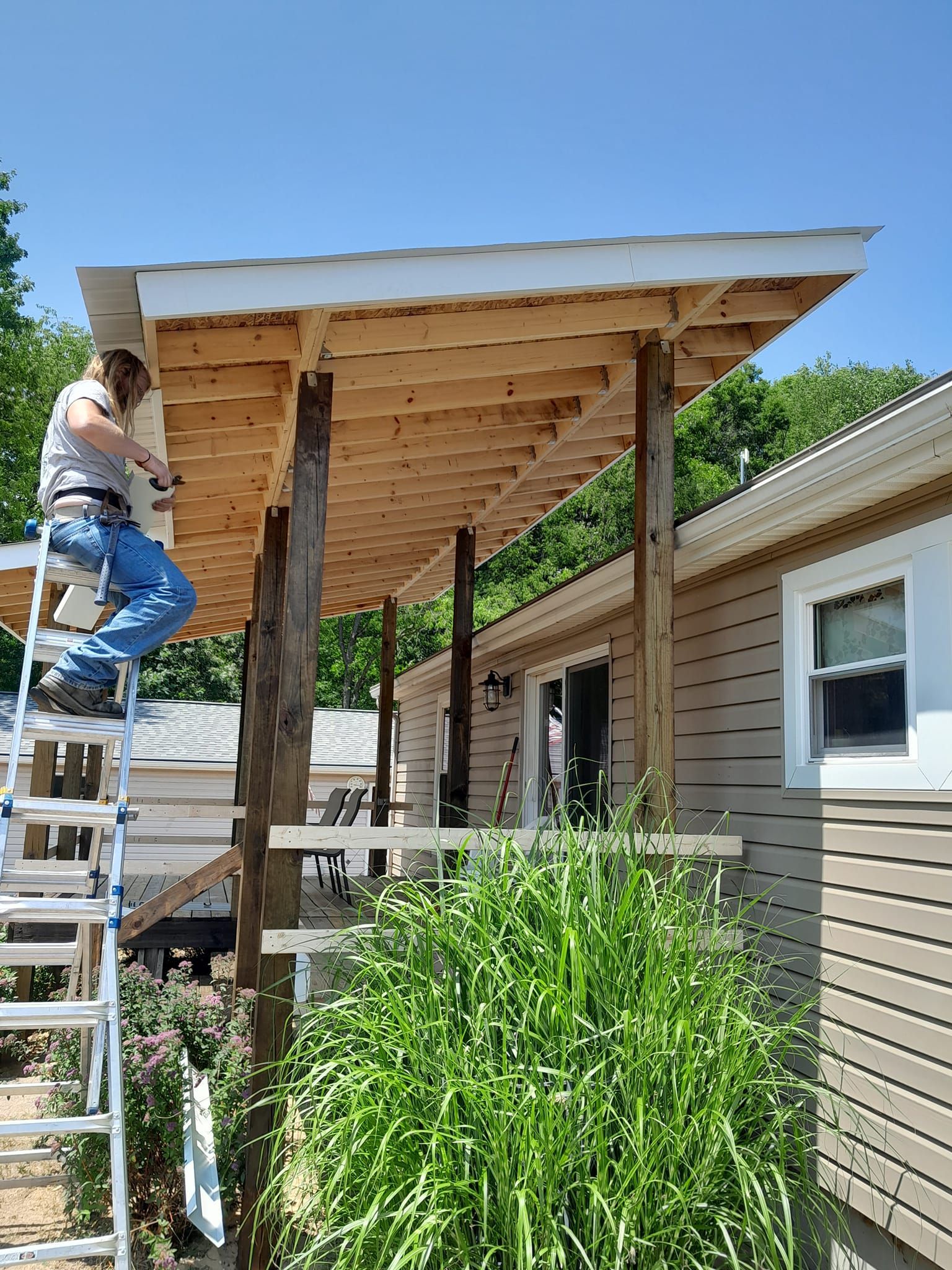 Person on a ladder building a wooden porch roof beside a house and greenhouse