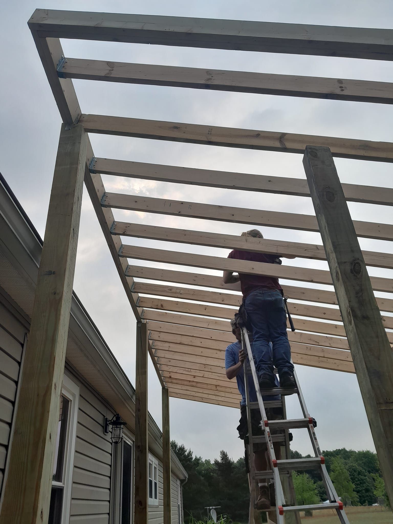 Worker on a ladder installing wooden beams on a pergola beside a house