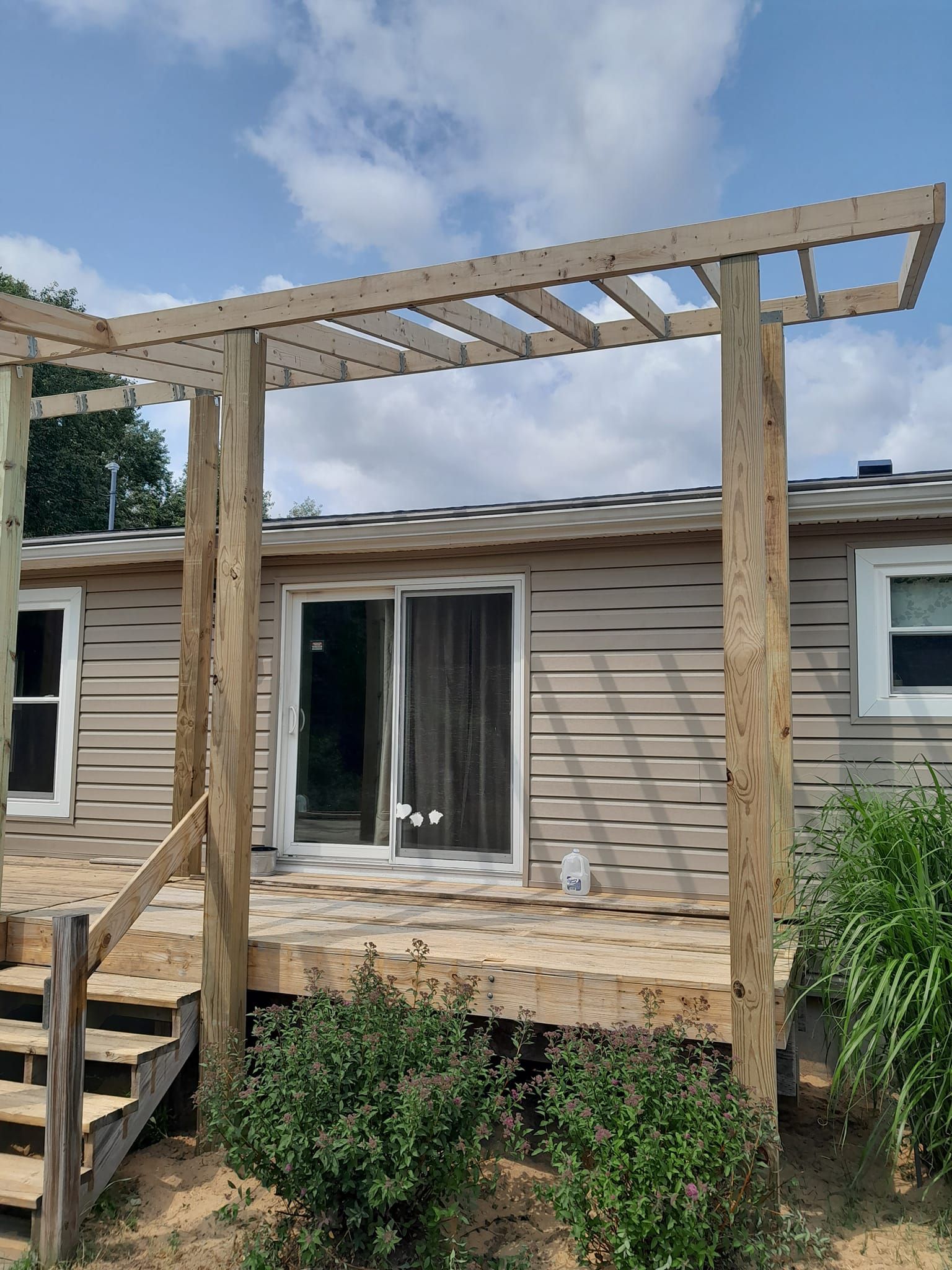 Backyard deck under construction with wooden pergola frame beside a beige house and sliding glass door