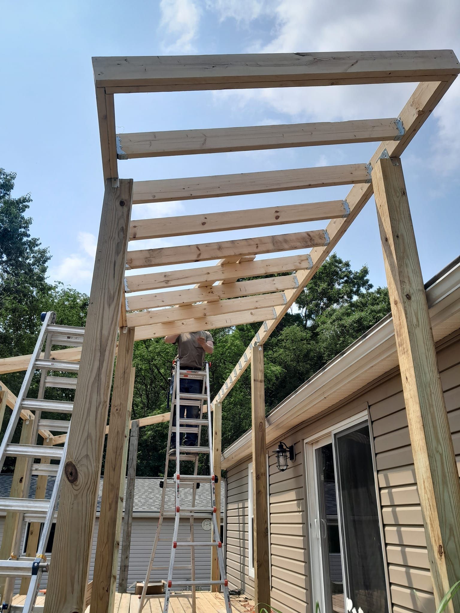 Wooden pergola frame under construction beside a house, with ladders and scaffolding visible.