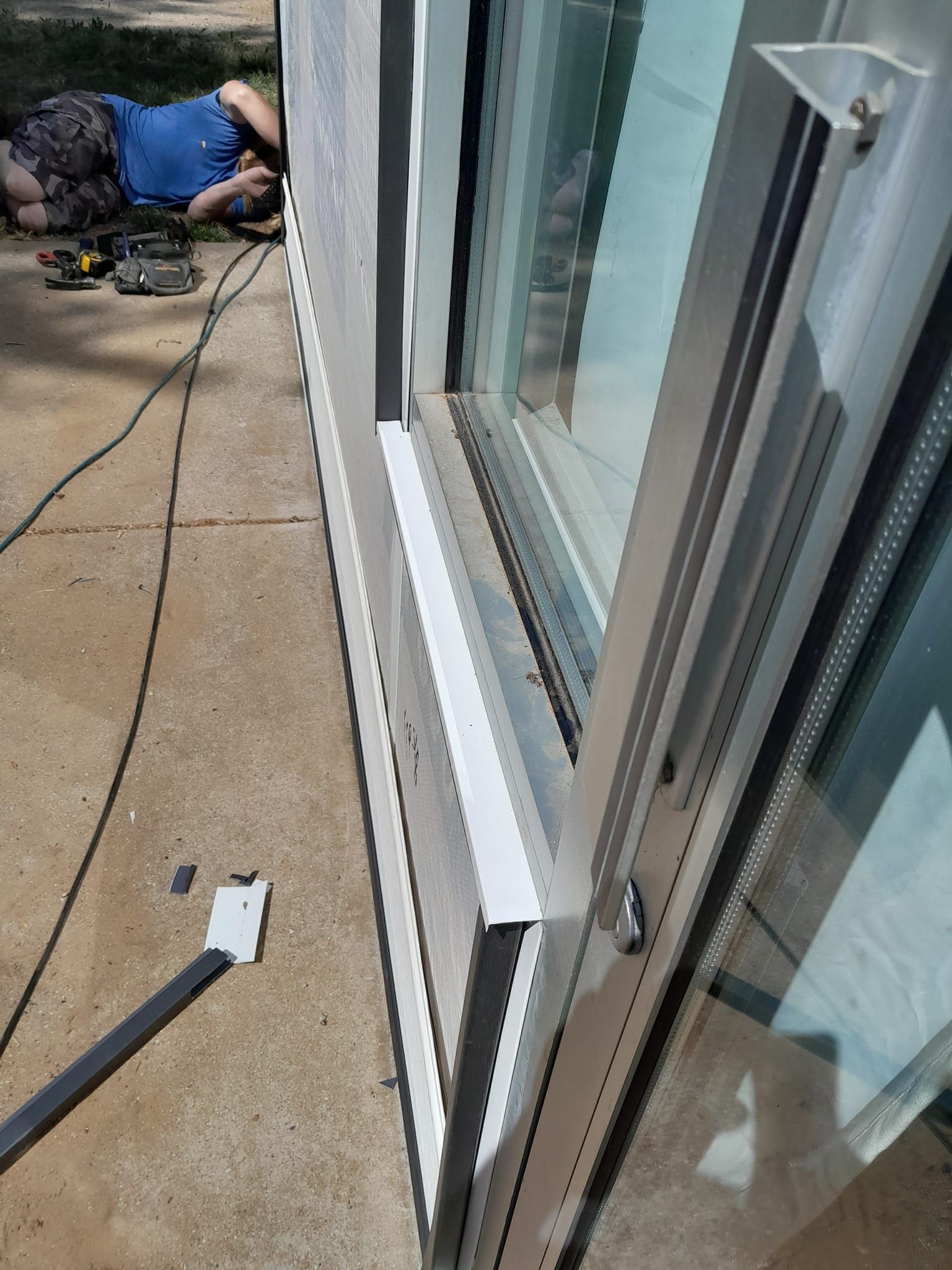 Worker on a concrete patio installing a white sliding glass door frame beside a house.