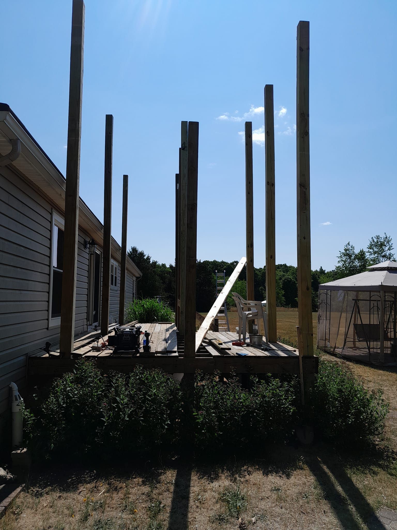 Partially built wooden house frame with tall posts beside a white house under a blue sky