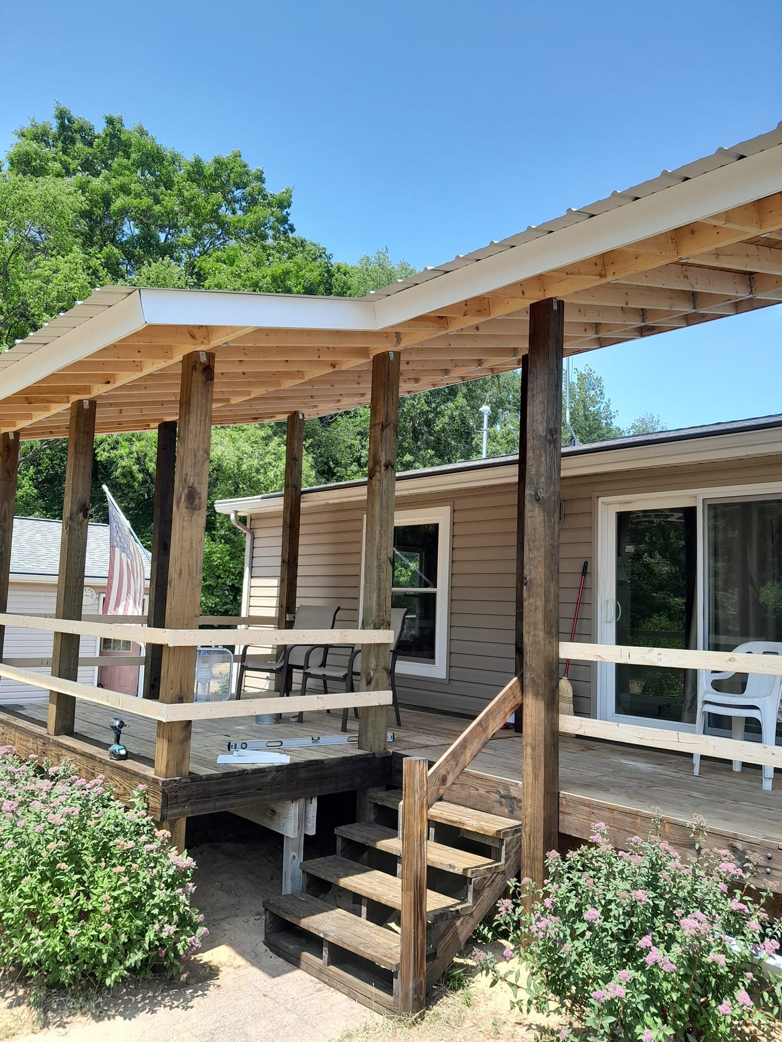 Wooden porch with stairs and pergola beside a tan house, with railings and outdoor seating in a sunny yard