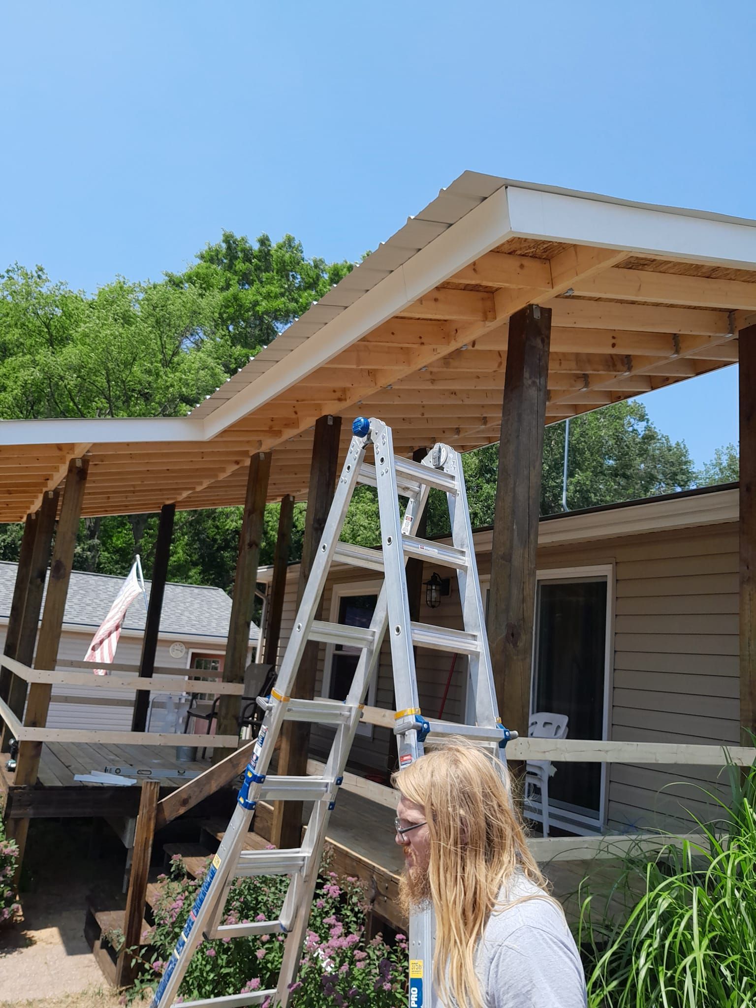Person on ladder under a wooden roof frame, working on a house porch in bright daylight