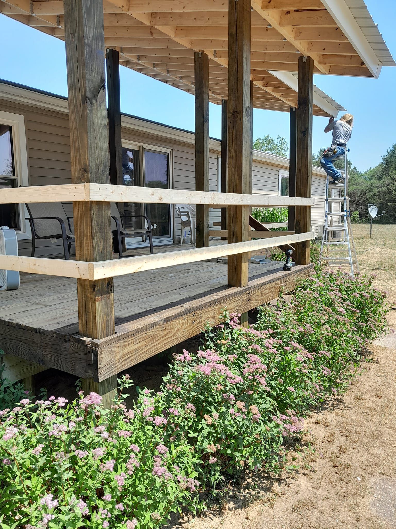 Wooden porch with white railings and pink flowering shrubs along a dry walkway under a blue sky