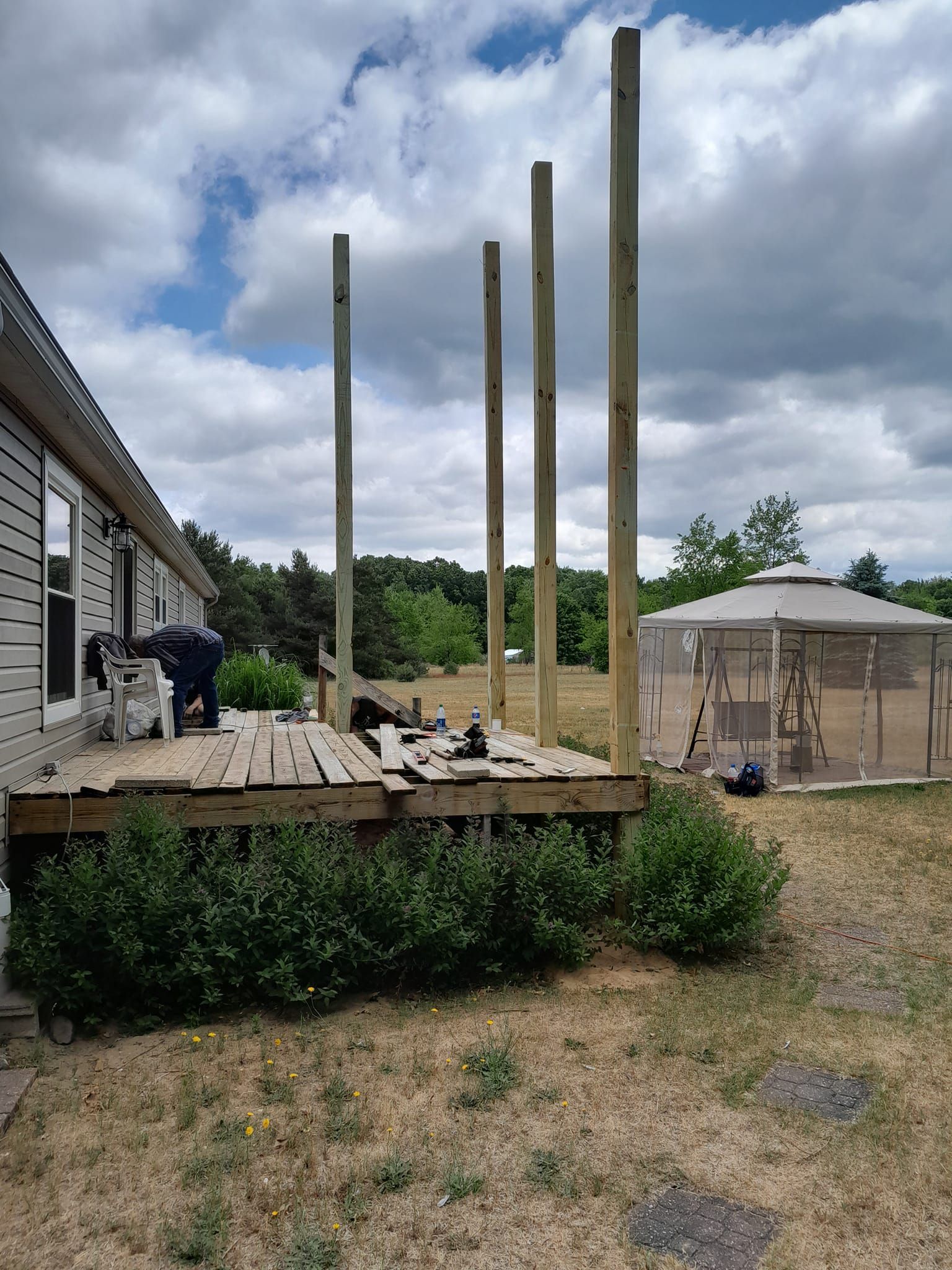 Backyard deck under construction with tall wooden posts beside a house and tan gazebo canopy