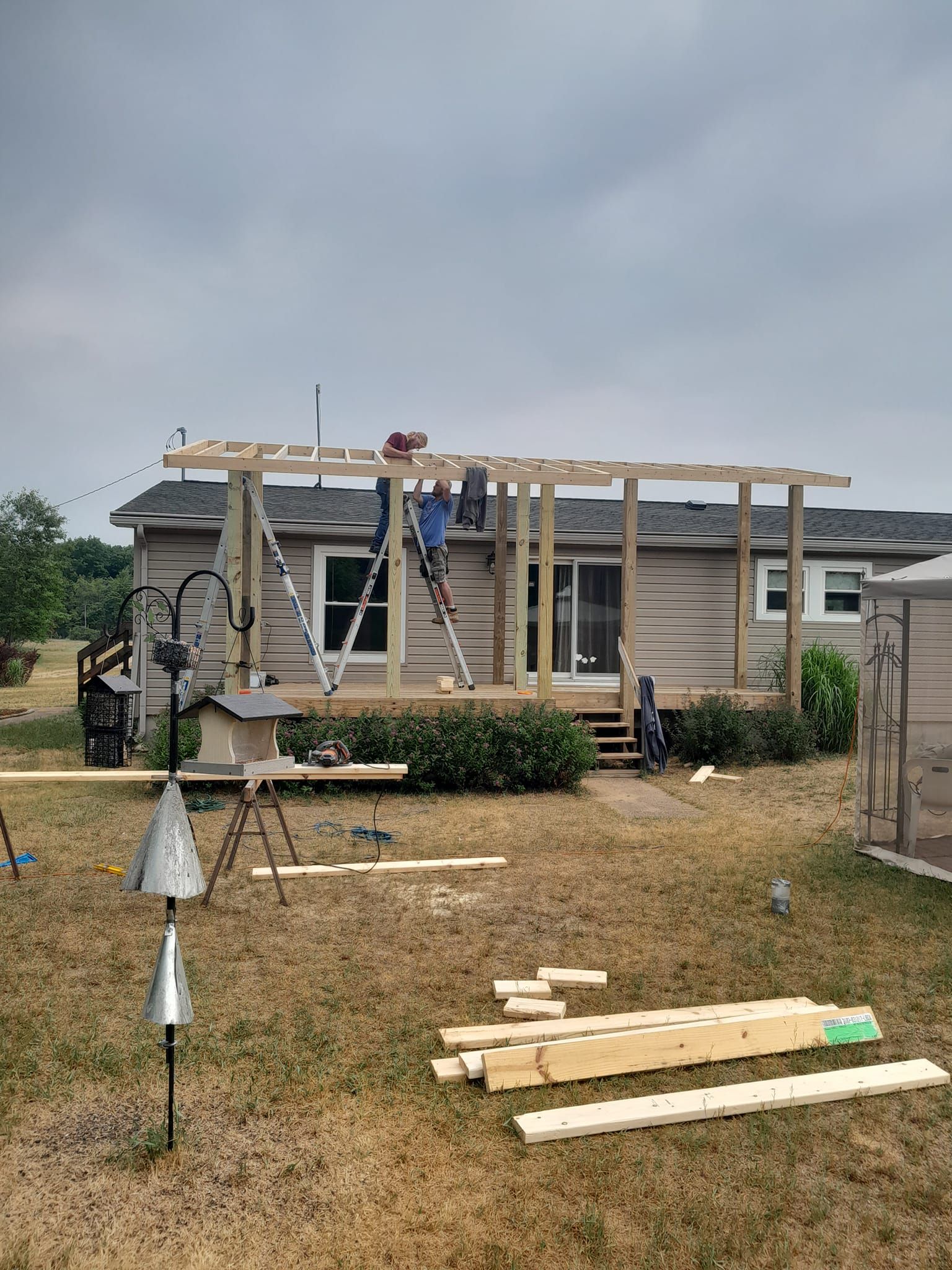 Partially built wooden structure with ladders and scattered lumber on a grassy lot under a cloudy sky