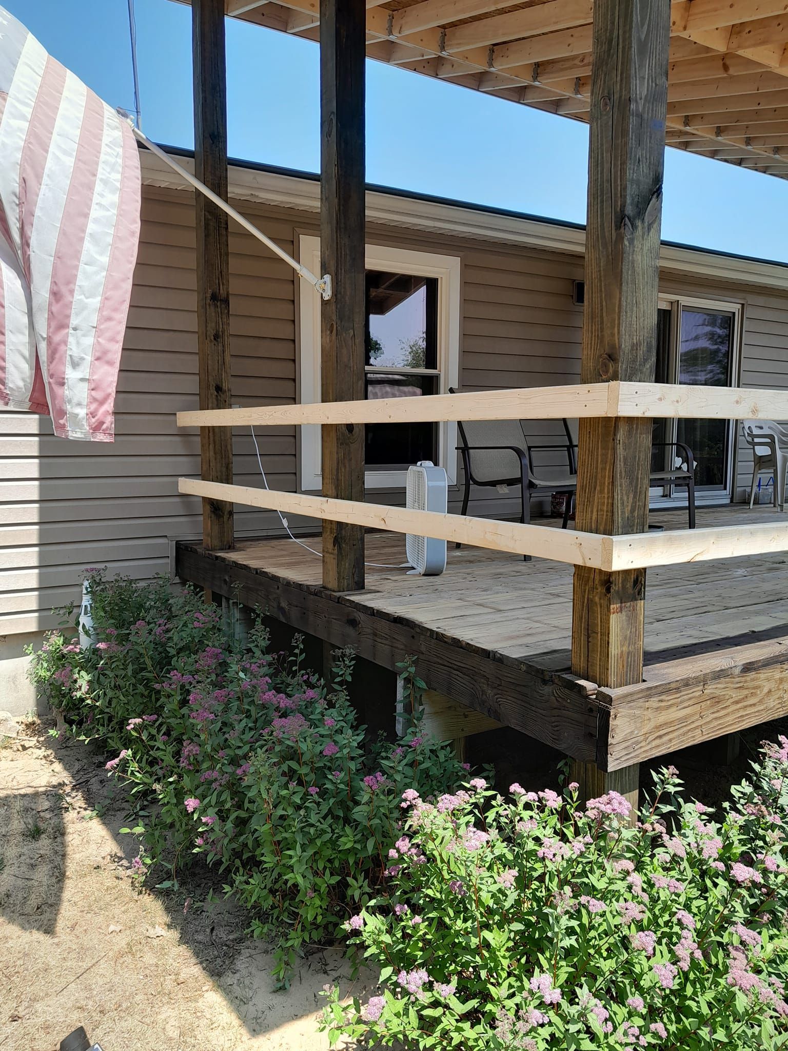 Wooden porch with railing and steps beside a beige house, with pink flowers in front.