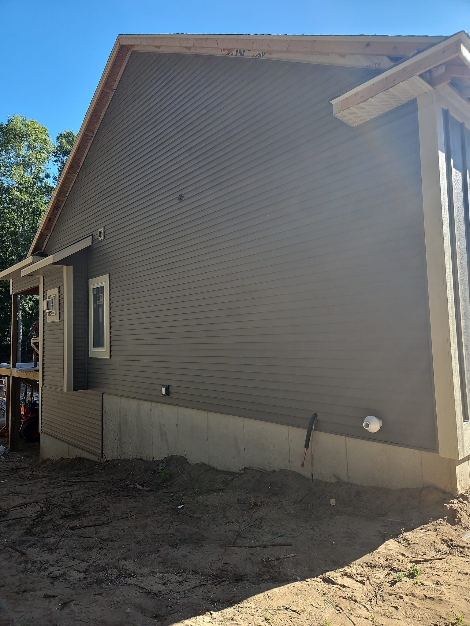 Exterior side of a tan house with beige foundation and a small window on a dirt lot under bright sunlight