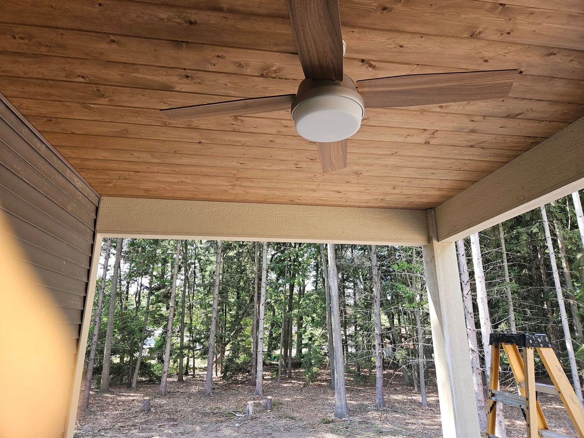 Covered porch with wood ceiling, ceiling fan, and wooded yard view through open sides
