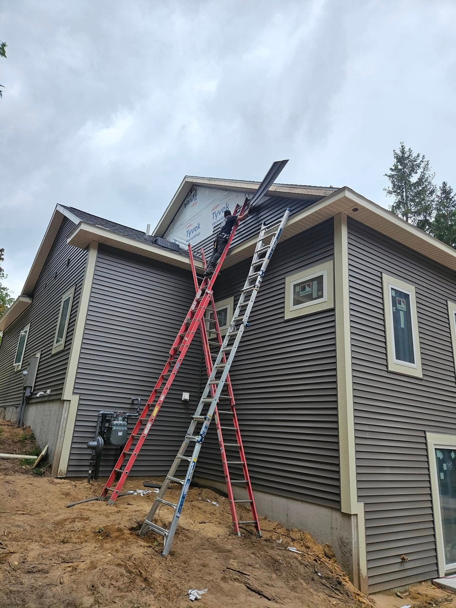 Ladders leaned against a house exterior with damaged roof shingles and siding repair underway.