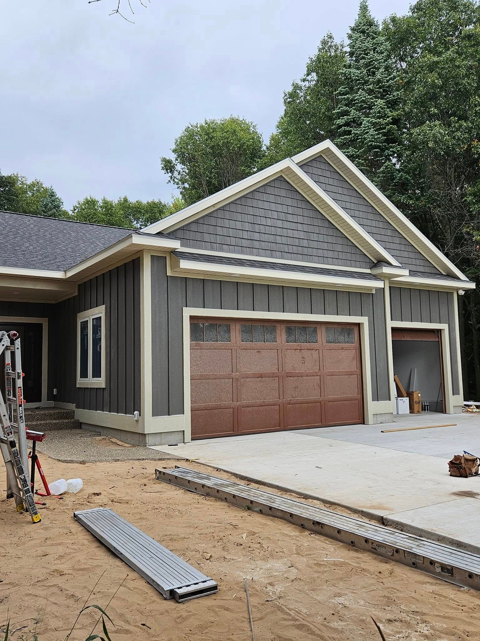 Front view of a house under construction with gray siding, a brown garage door, and unfinished driveway and roof.