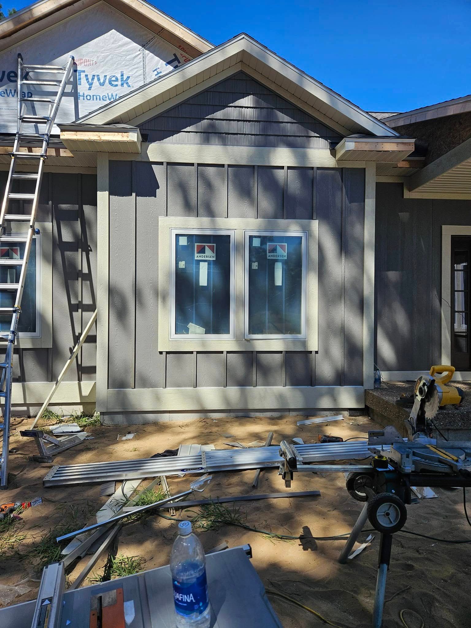 Gray house exterior under construction with ladders, windows, tools, and scattered building materials