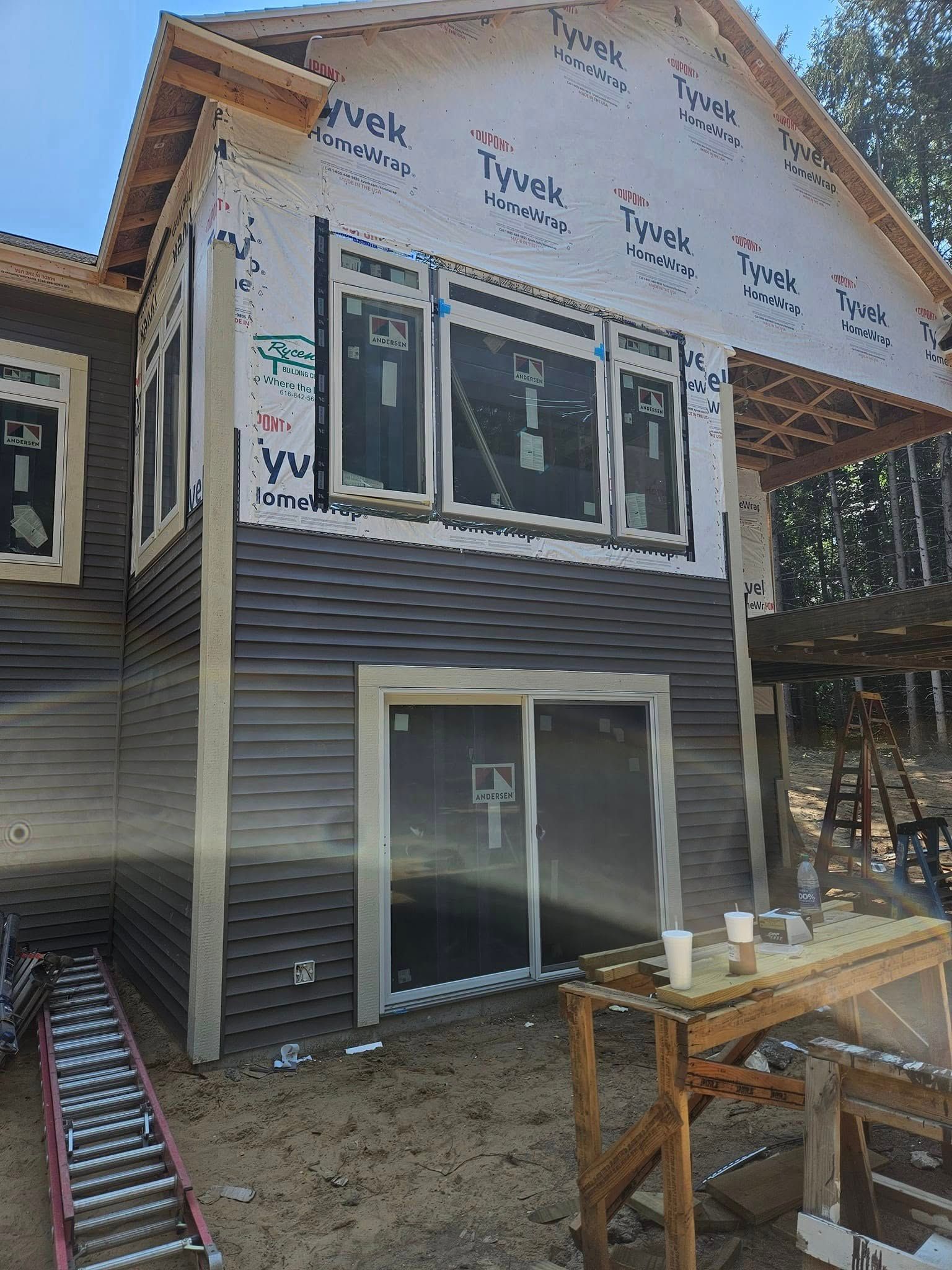 Two-story house under construction with gray siding, exposed roof sheathing, and scaffolding nearby