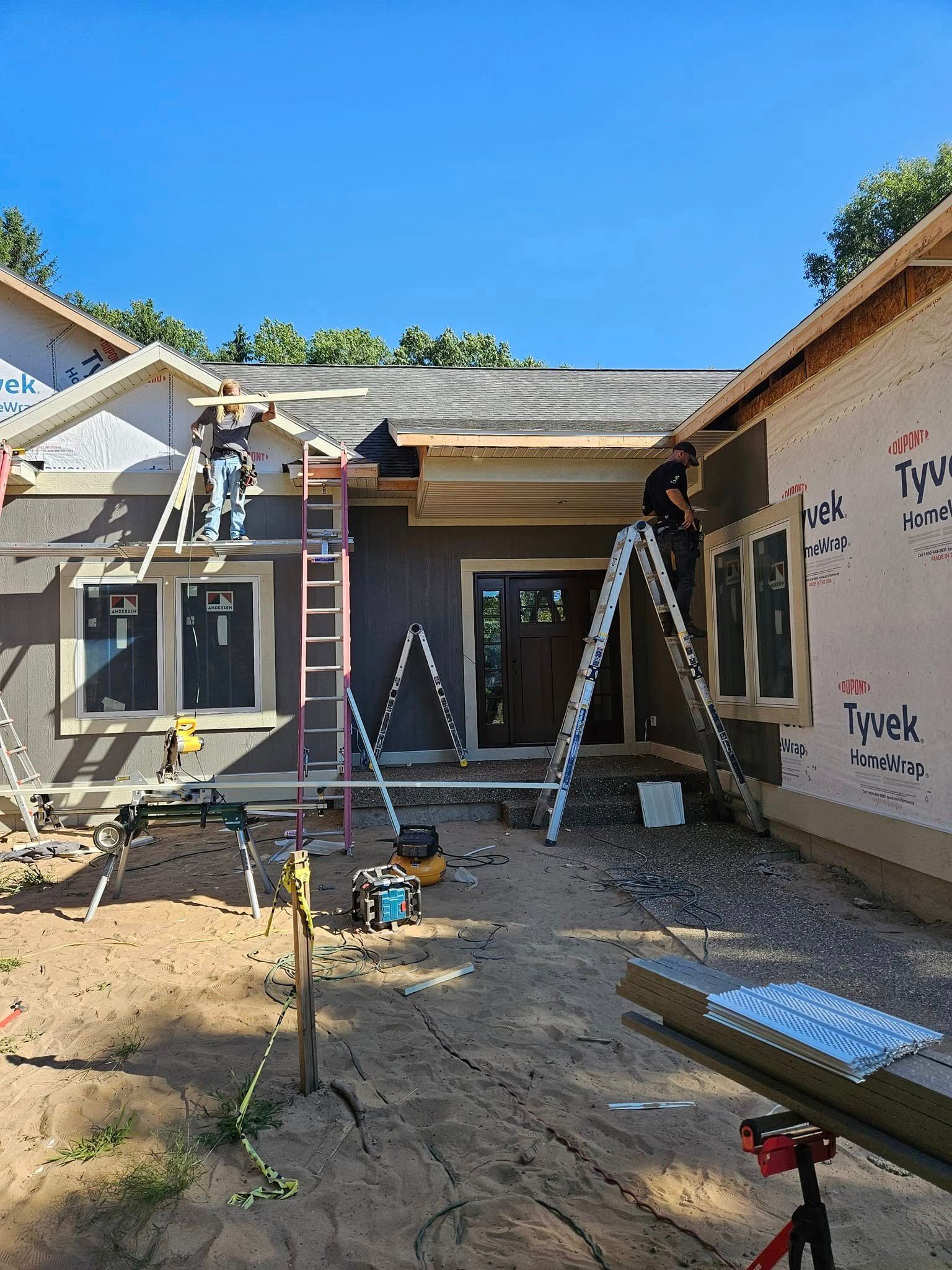 Front entrance of a house under construction, with ladders, scaffolding, and workers on the roof.