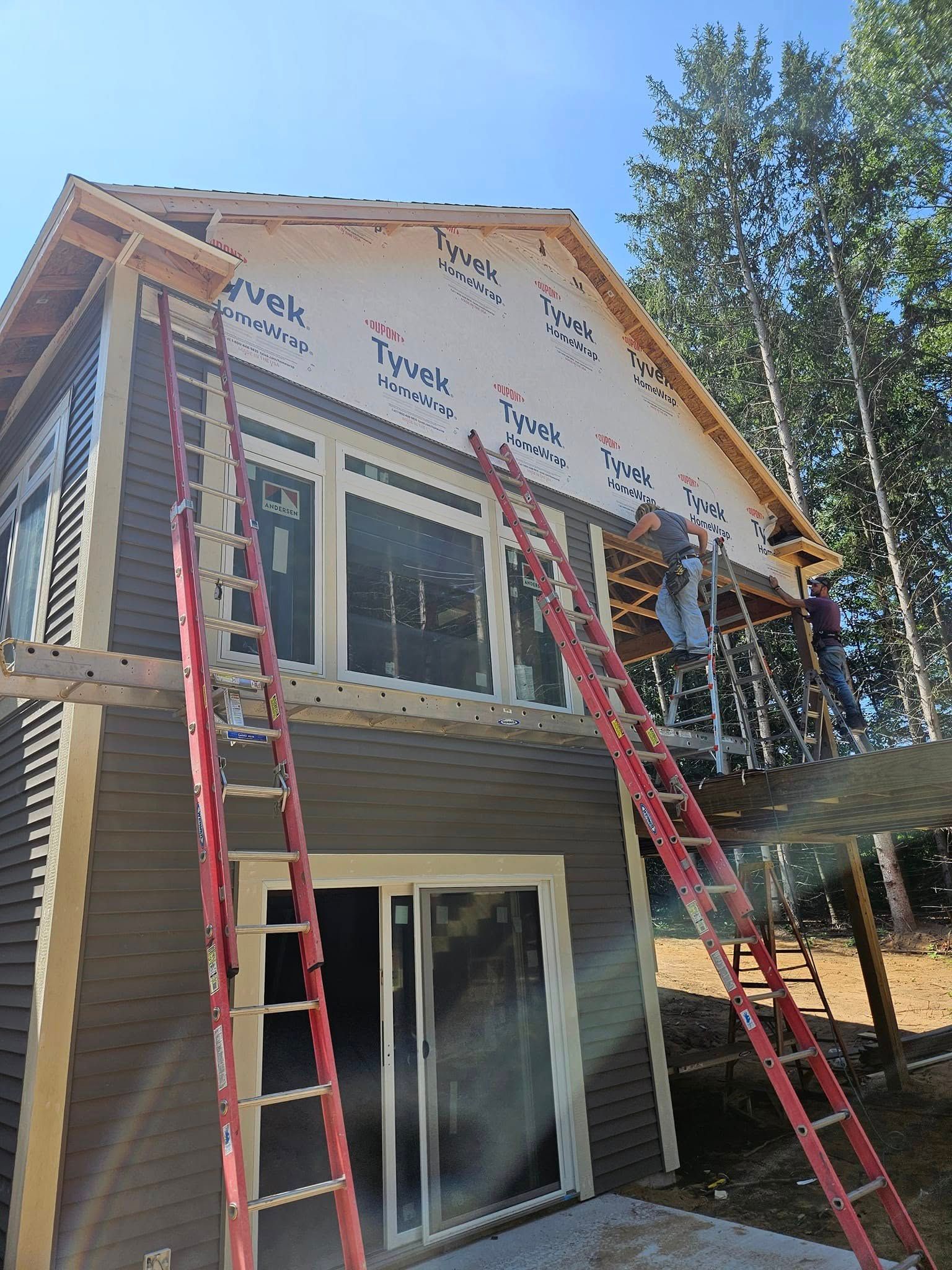 House exterior under construction with two red ladders and exposed roof sheathing above sliding glass doors