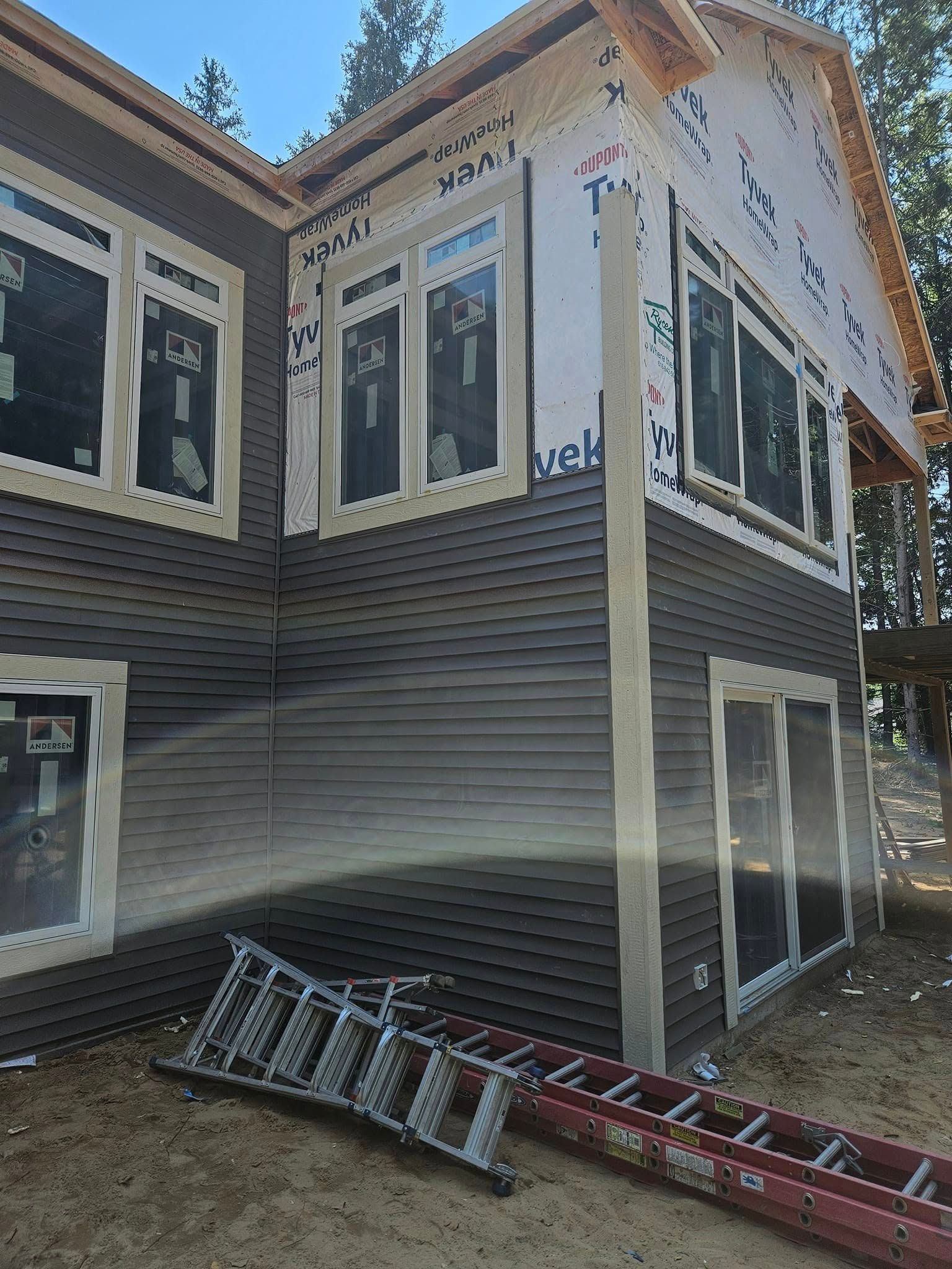 Two-story house under construction with dark siding, white trim, and ladders leaning against it