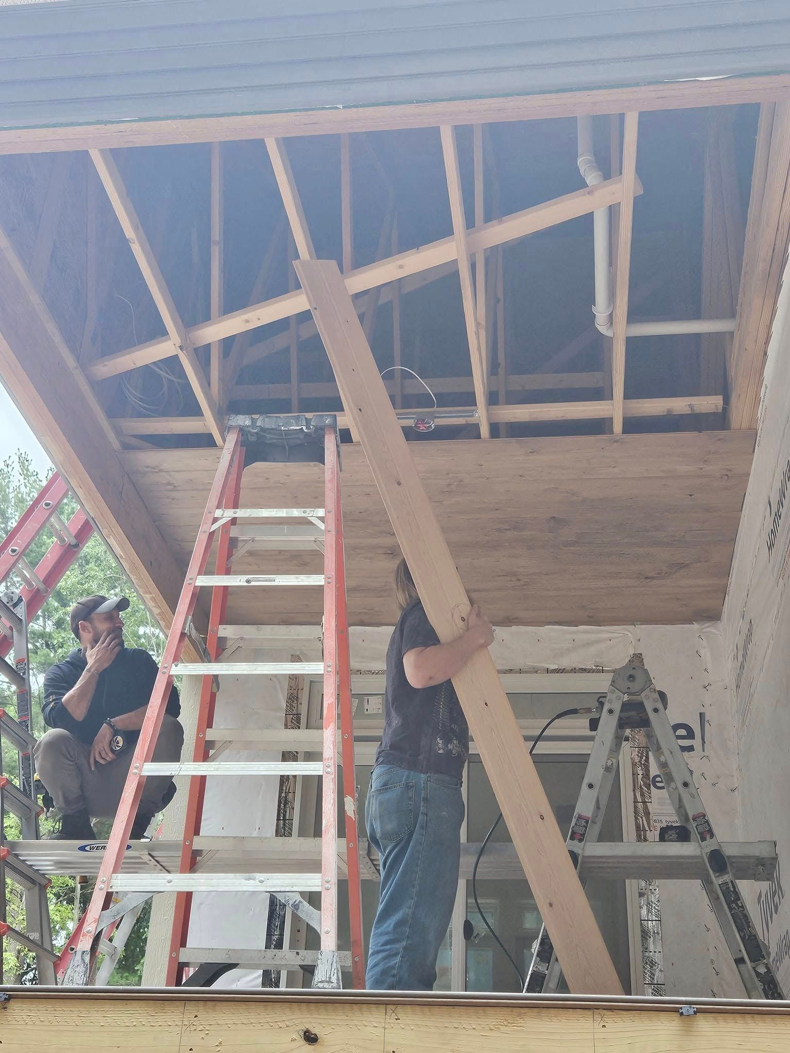 People on ladders framing a ceiling with plywood and exposed rafters at a construction site