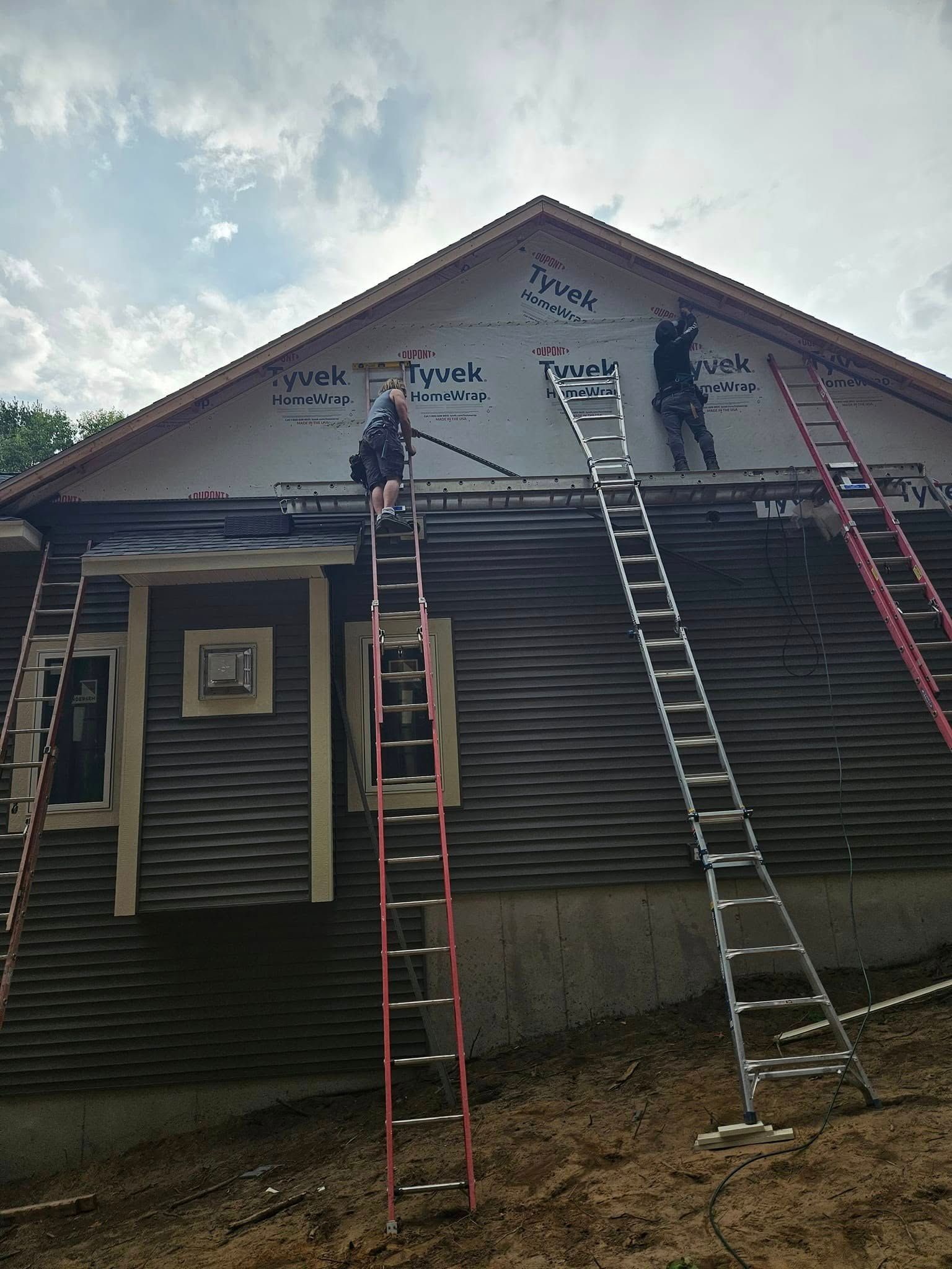 Workers on ladders repairing a house roof and siding under a cloudy sky