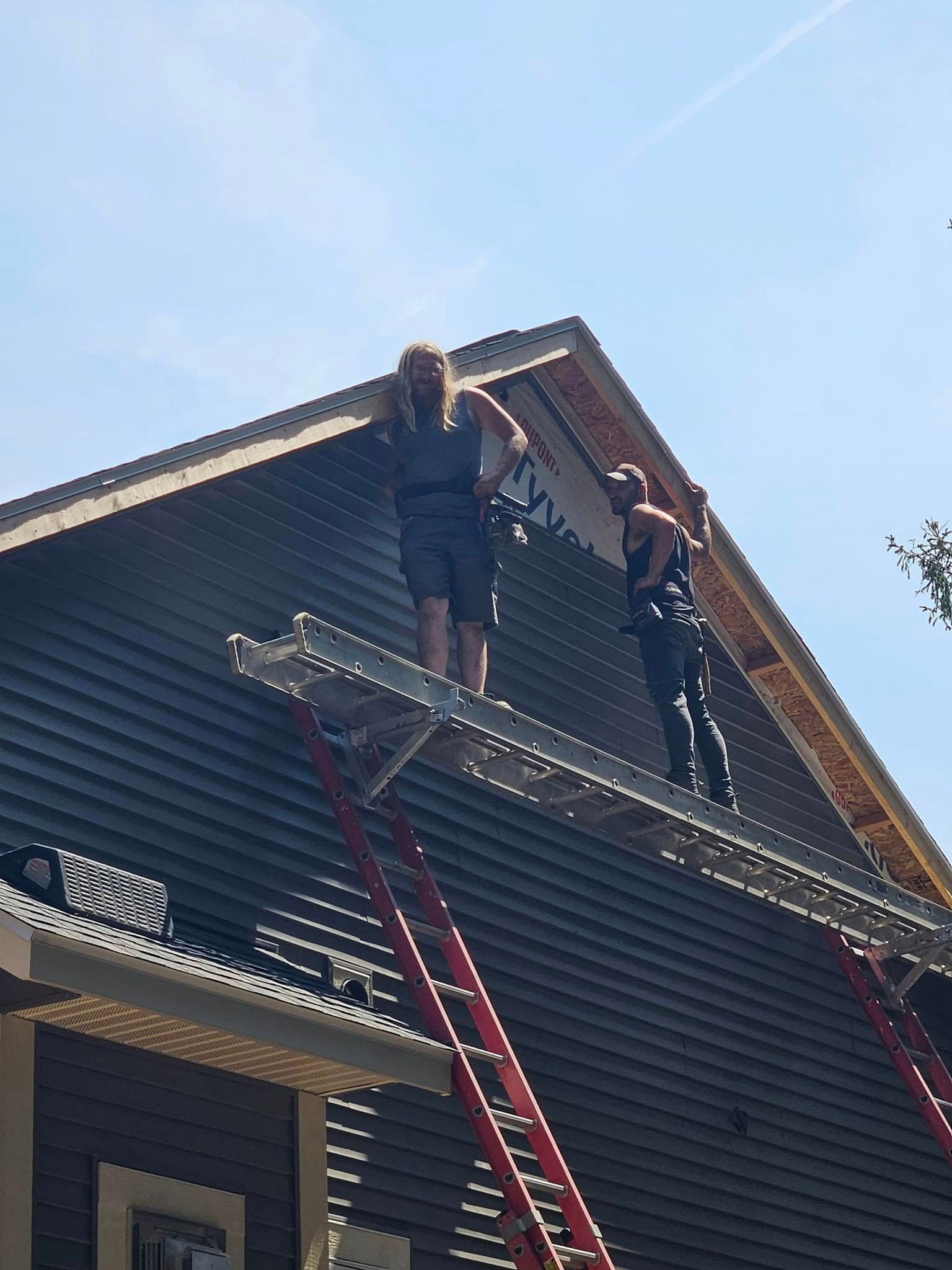 Two workers on a ladder installing siding on a house gable under a blue sky