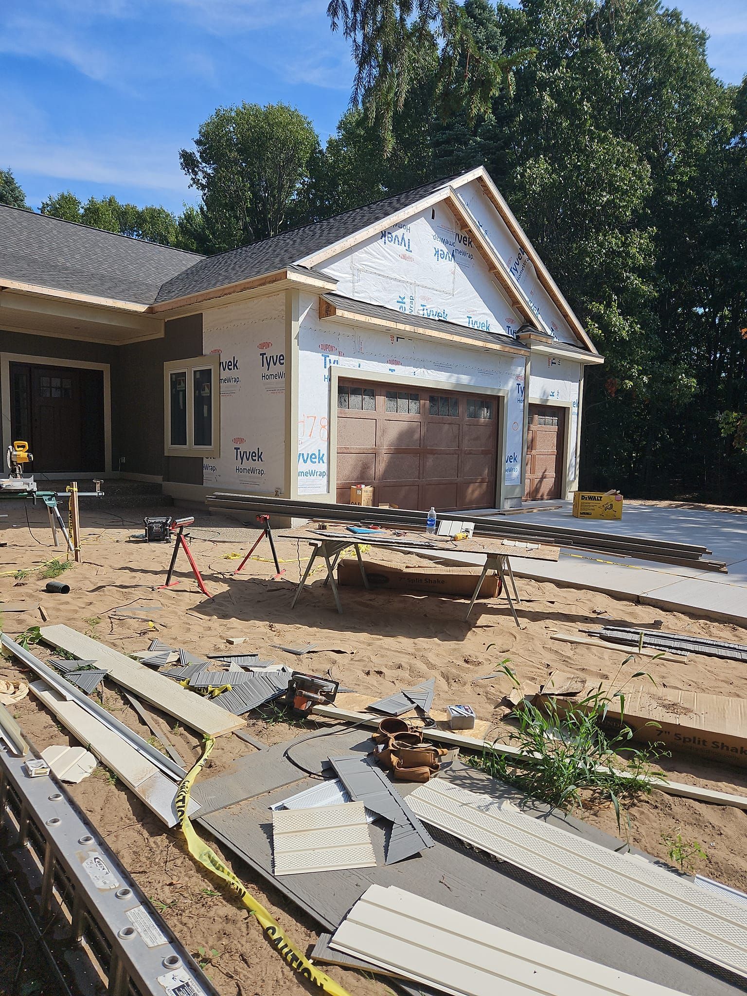 Partially built house with detached garage, construction debris, and dirt yard under a sunny blue sky