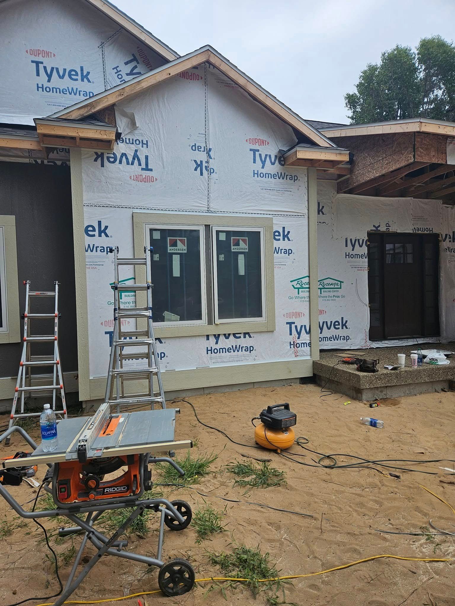 Front of a house under construction with ladders, tools, and unfinished siding in a muddy yard