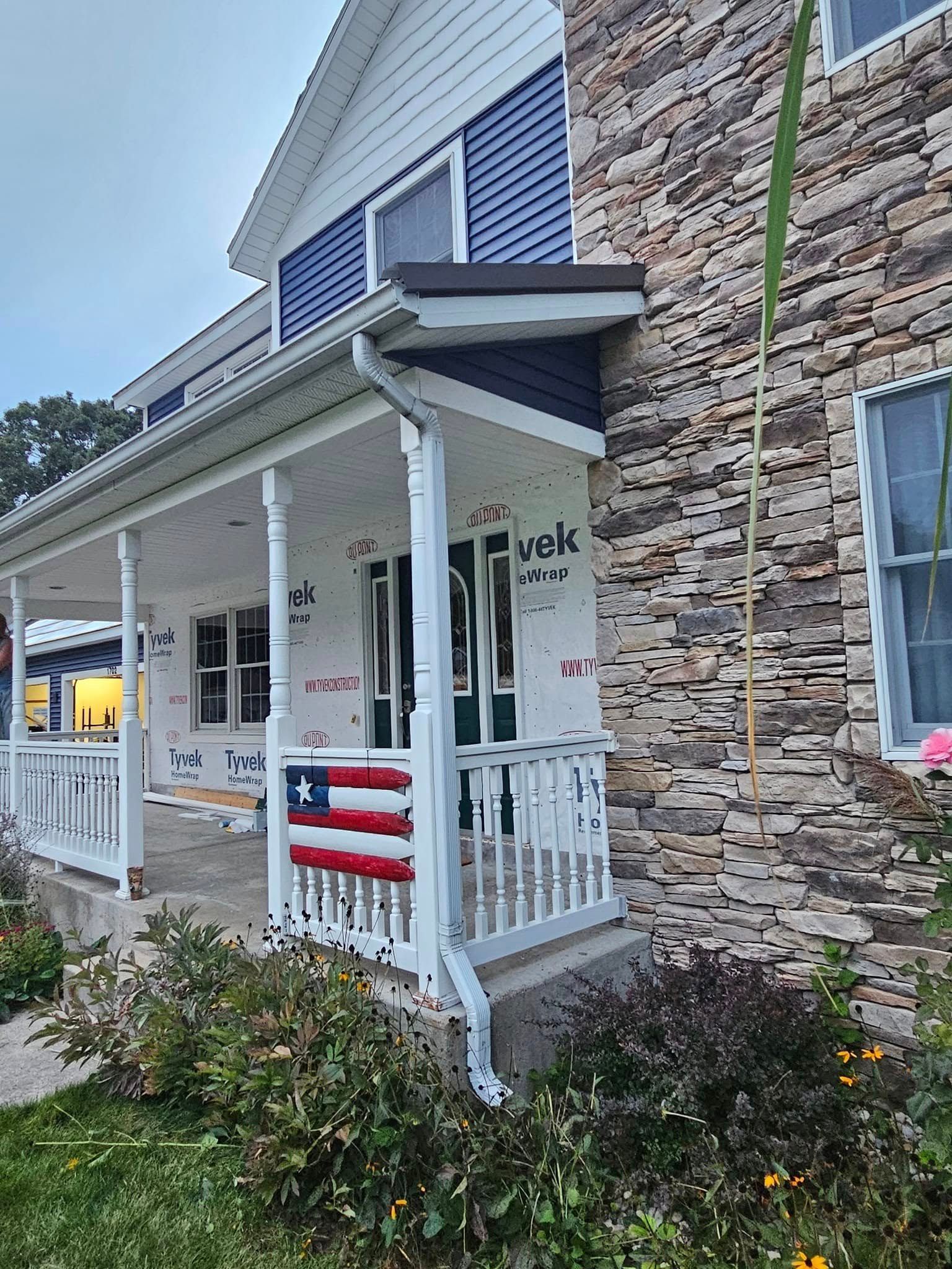Covered porch with white railings, stone and blue siding, and an American flag draped over the front porch.