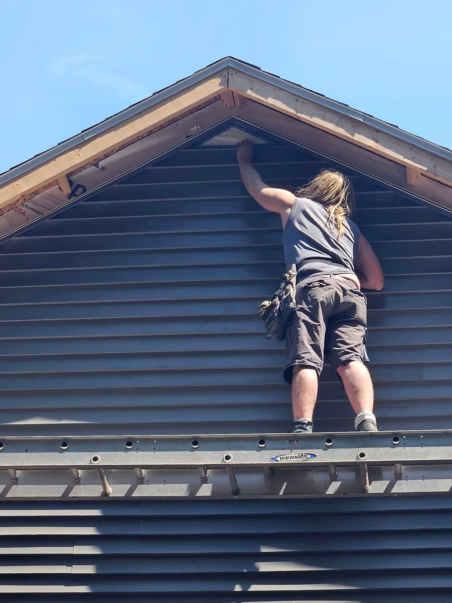 Person standing on a roof, reaching up to repair the eaves under a blue sky.