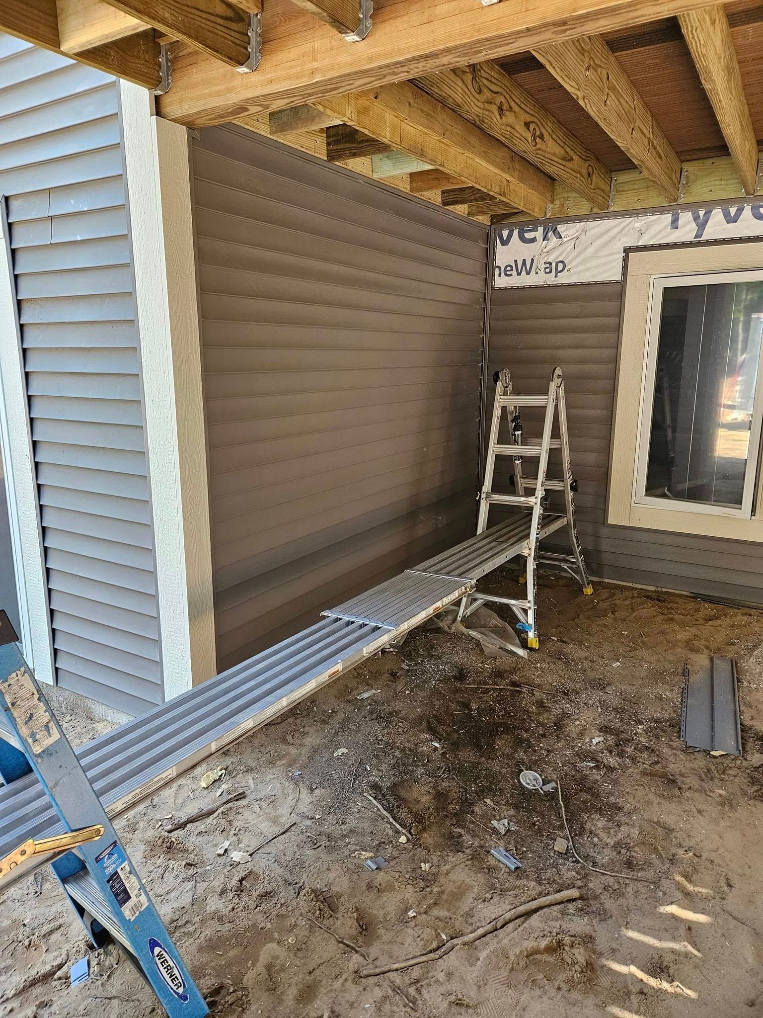 Worker on ladder under porch roof, installing brown siding on a house exterior.