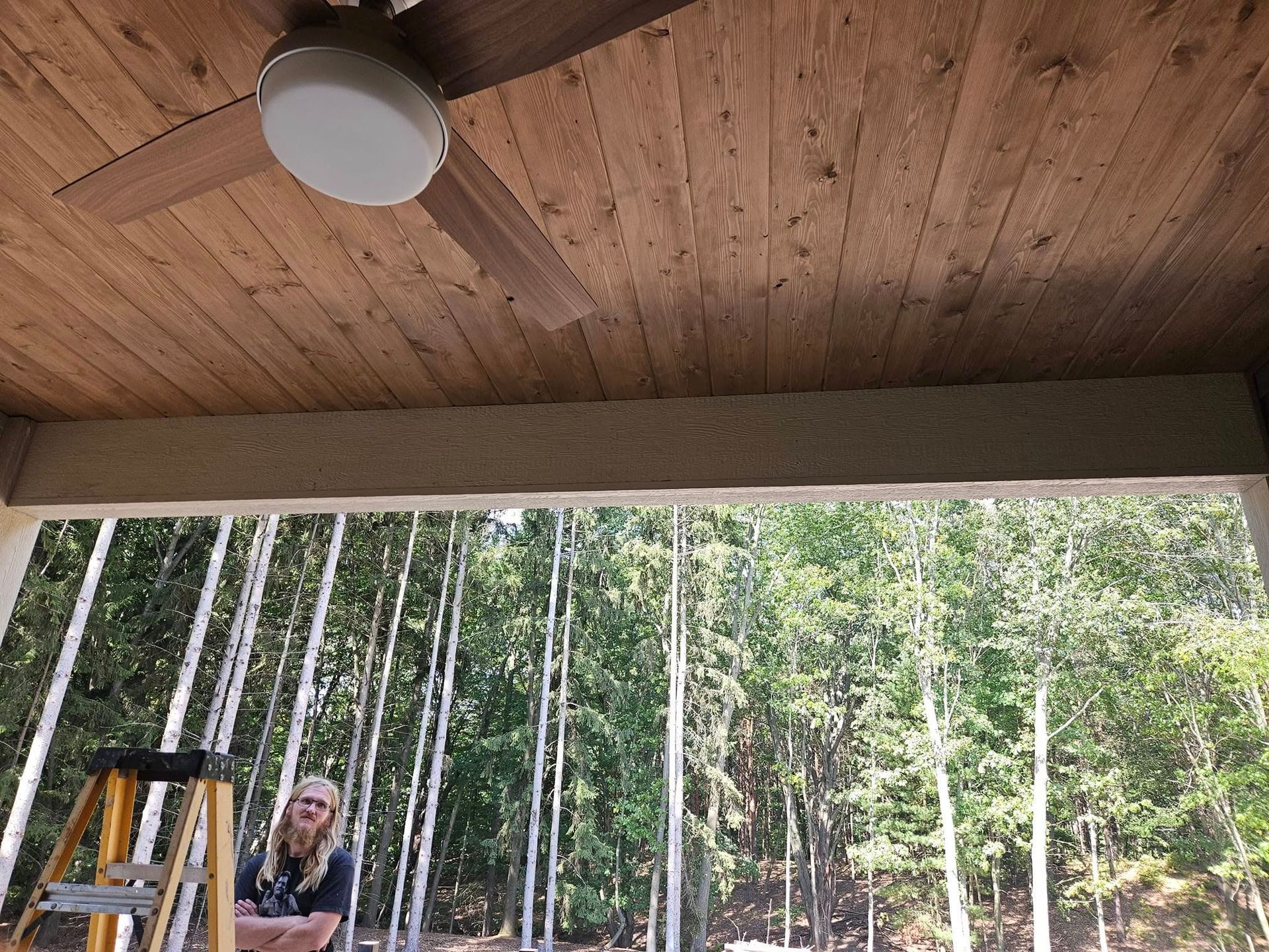 Wooden porch ceiling with ceiling fan, overlooking a forest; a person stands near a ladder below.