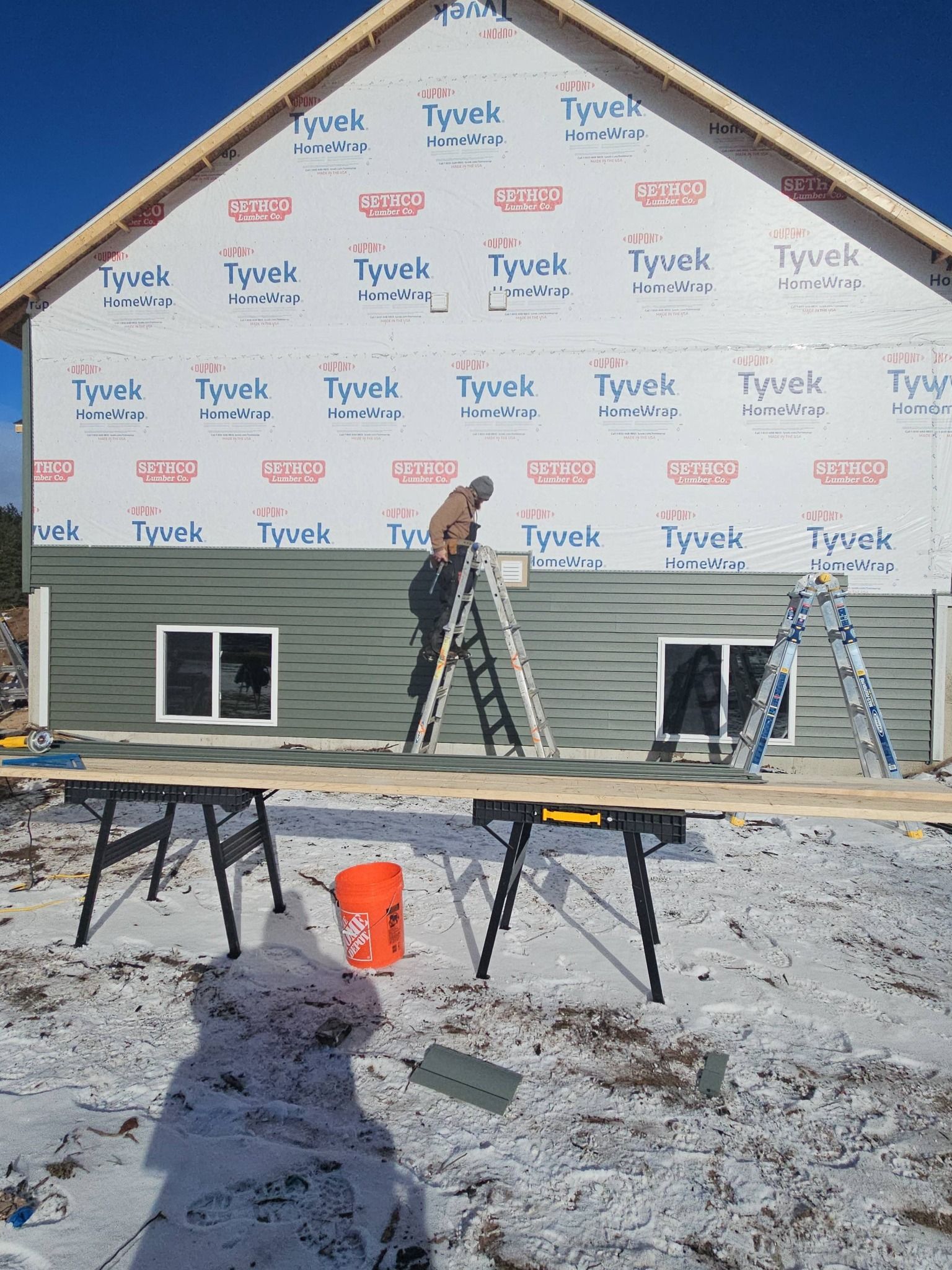 House under construction with white Tyvek wrap, two ladders, sawhorses, and snow-covered ground.