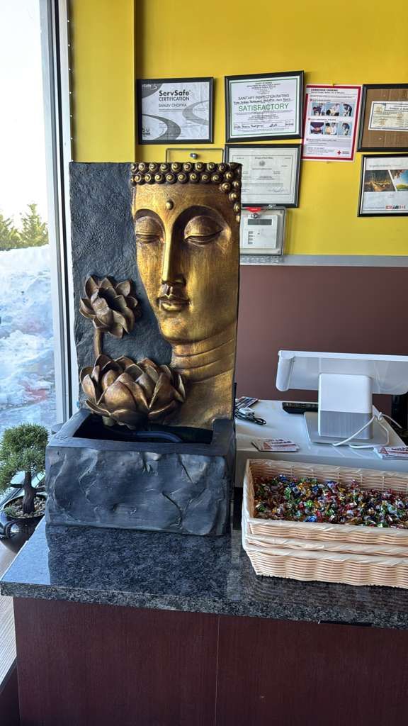 Buddha fountain with gold face, two lotus flowers, on a countertop, near a basket of colorful stones.