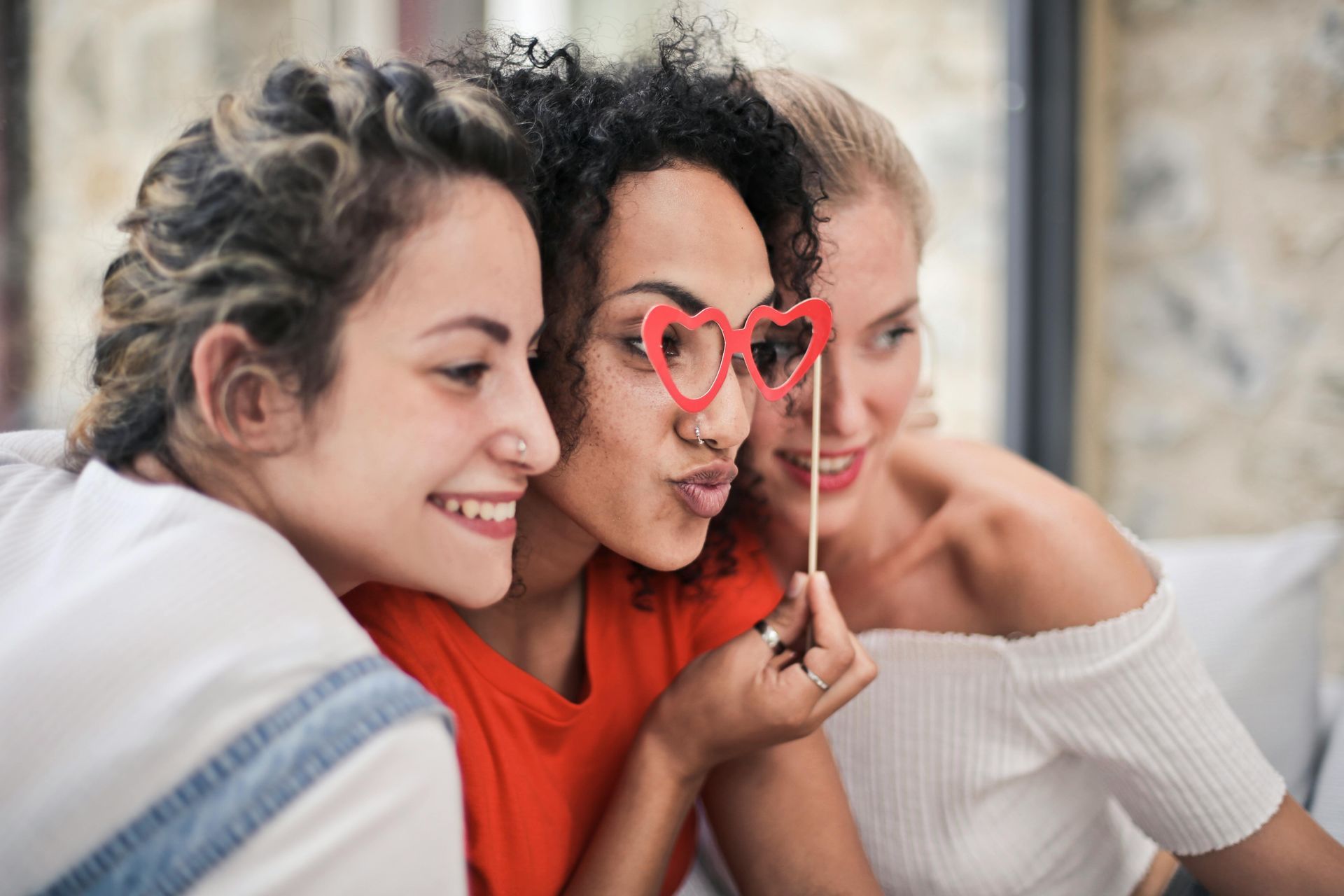 Three women smile while posing for a photo, one holding heart-shaped glasses.