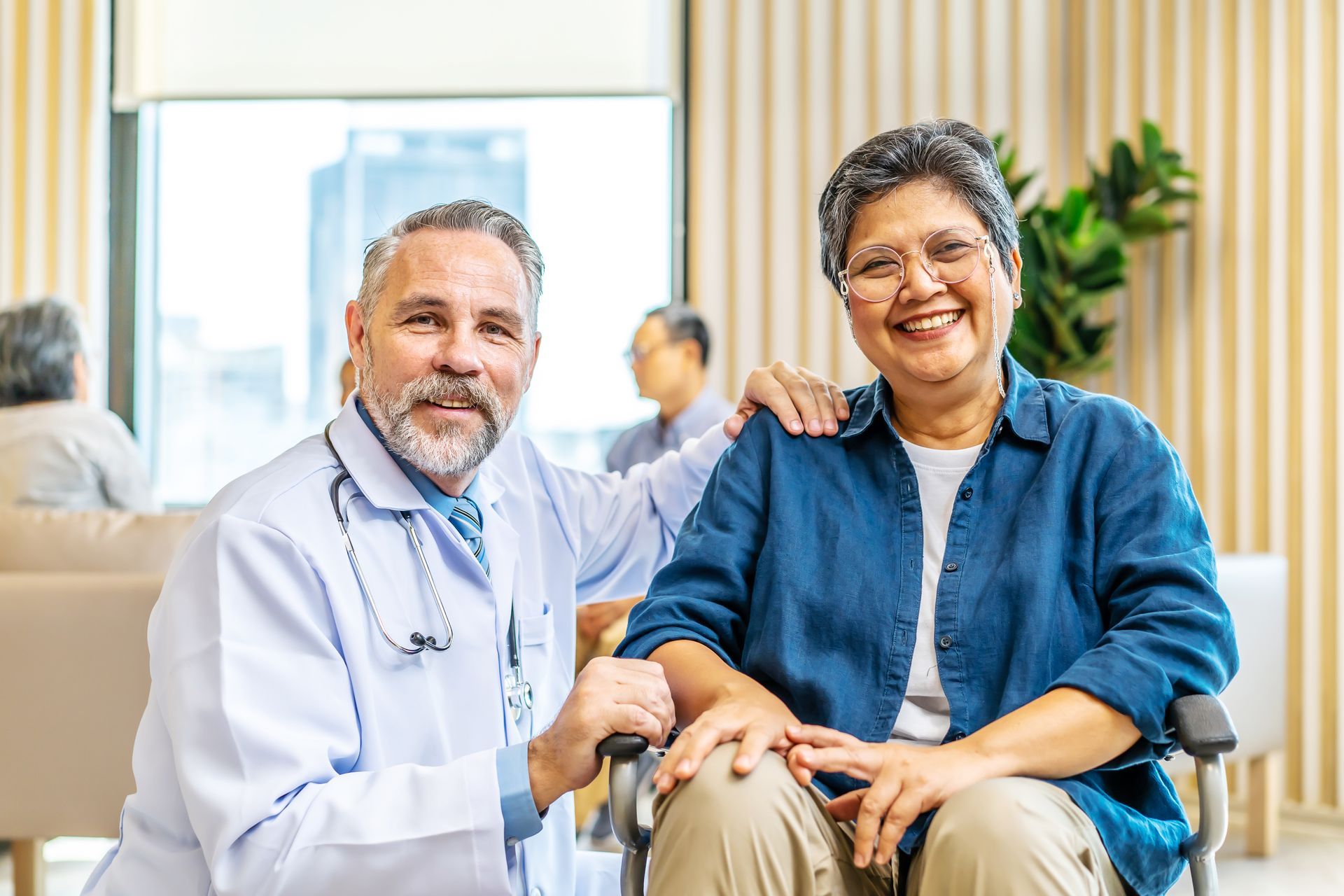 Doctor with stethoscope, arm around smiling woman in wheelchair; bright room.